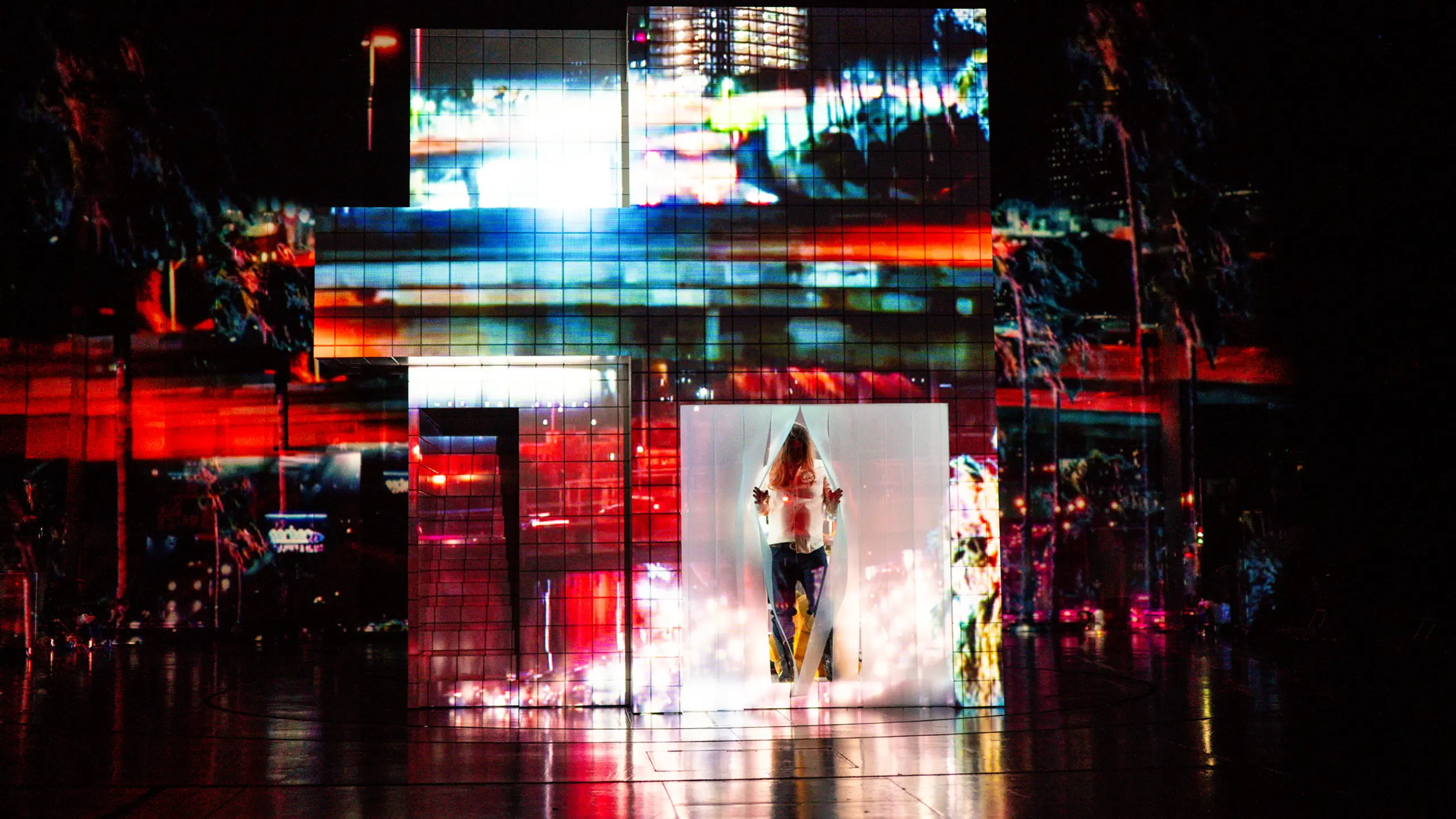 Skuespiller inne i transparent kube med projeksjoner av fargerike, urbane lys på fasadeformet struktur.