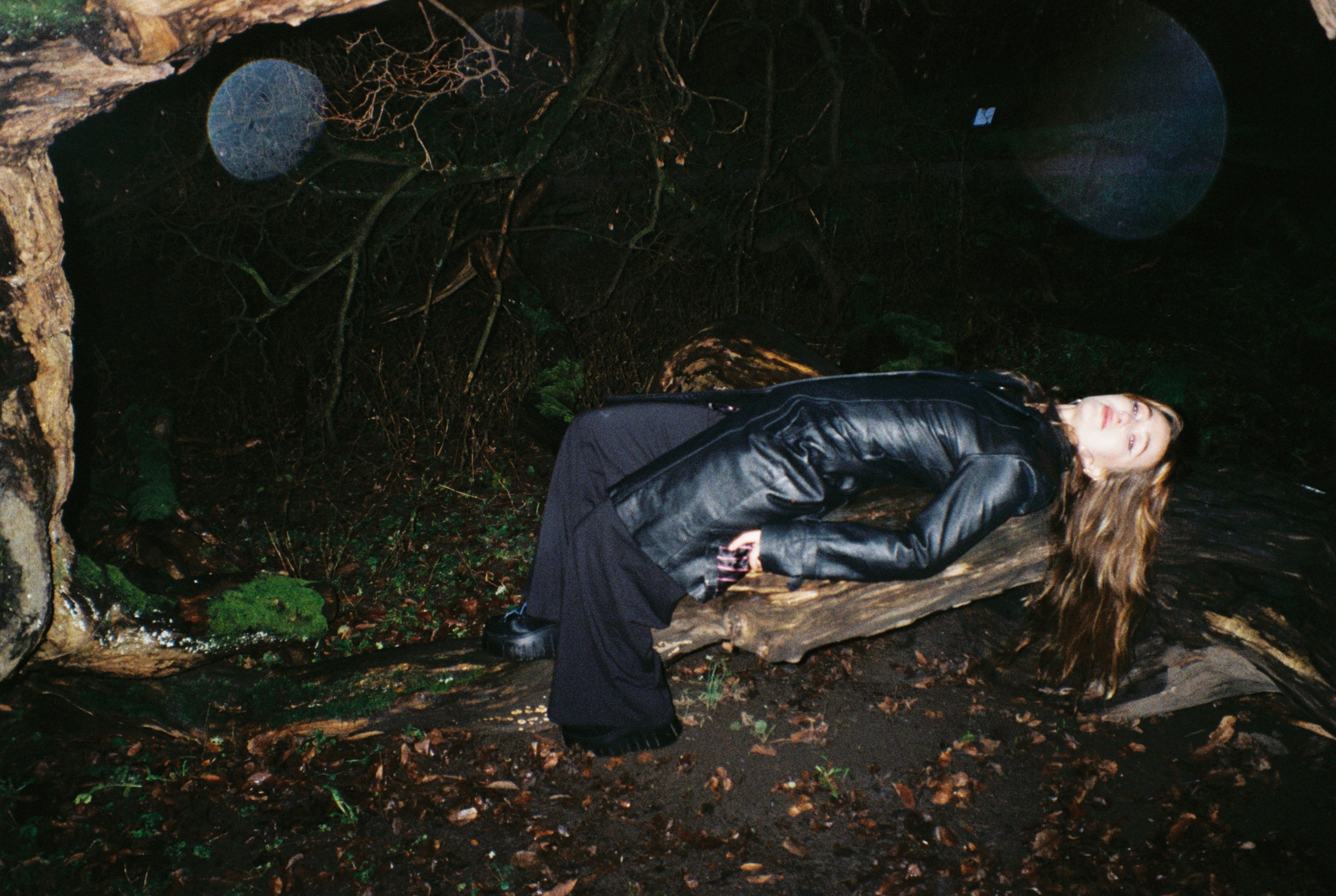 En ung kvinne i sort skinnfrakk ligger på en rot i mørket