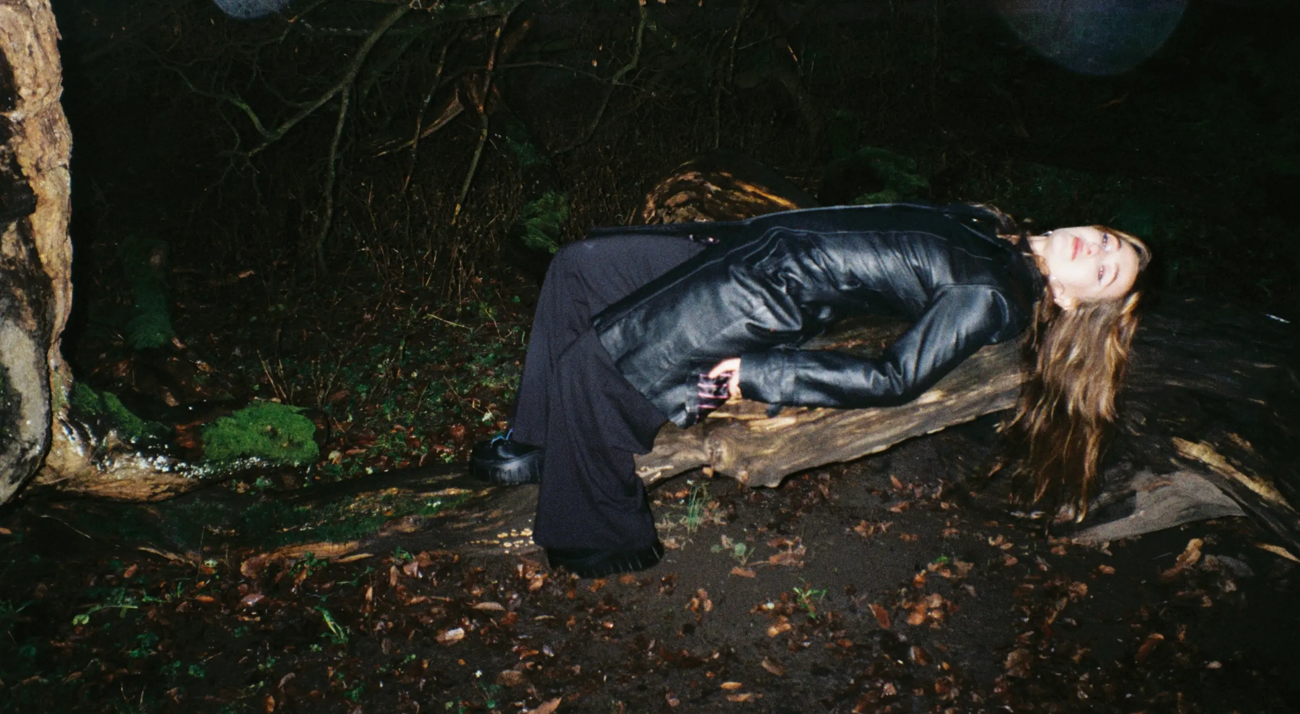 En ung kvinne i sort skinnfrakk ligger på en rot i mørket