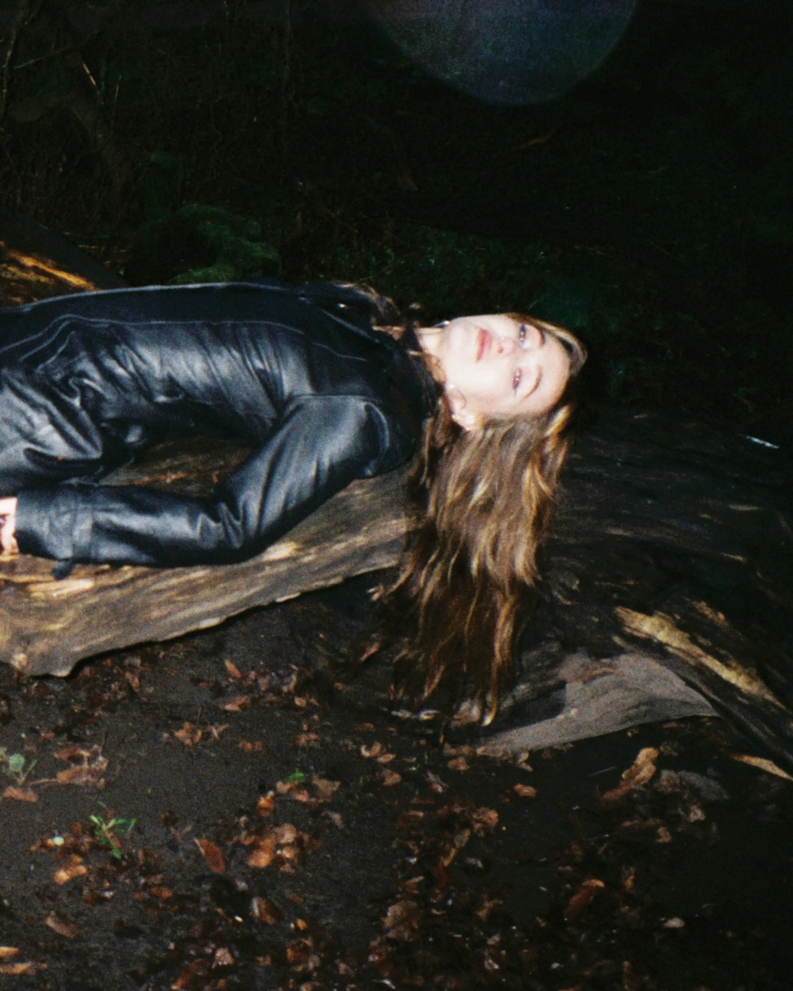 En ung kvinne i sort skinnfrakk ligger på en rot i mørket