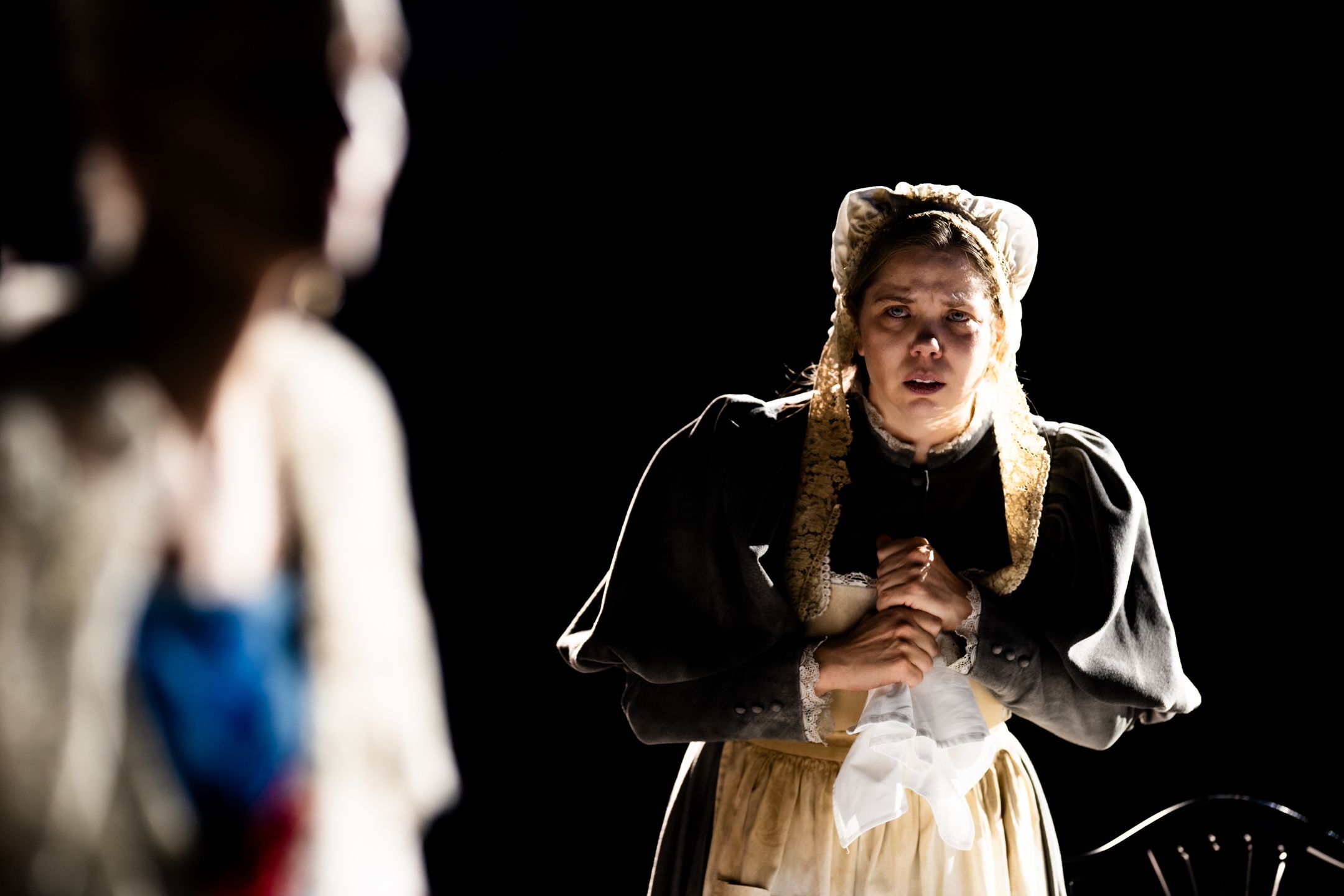 Kvinne i gammeldags kjole og kyse står med et hvitt lommetørkle og ser bekymret ut.