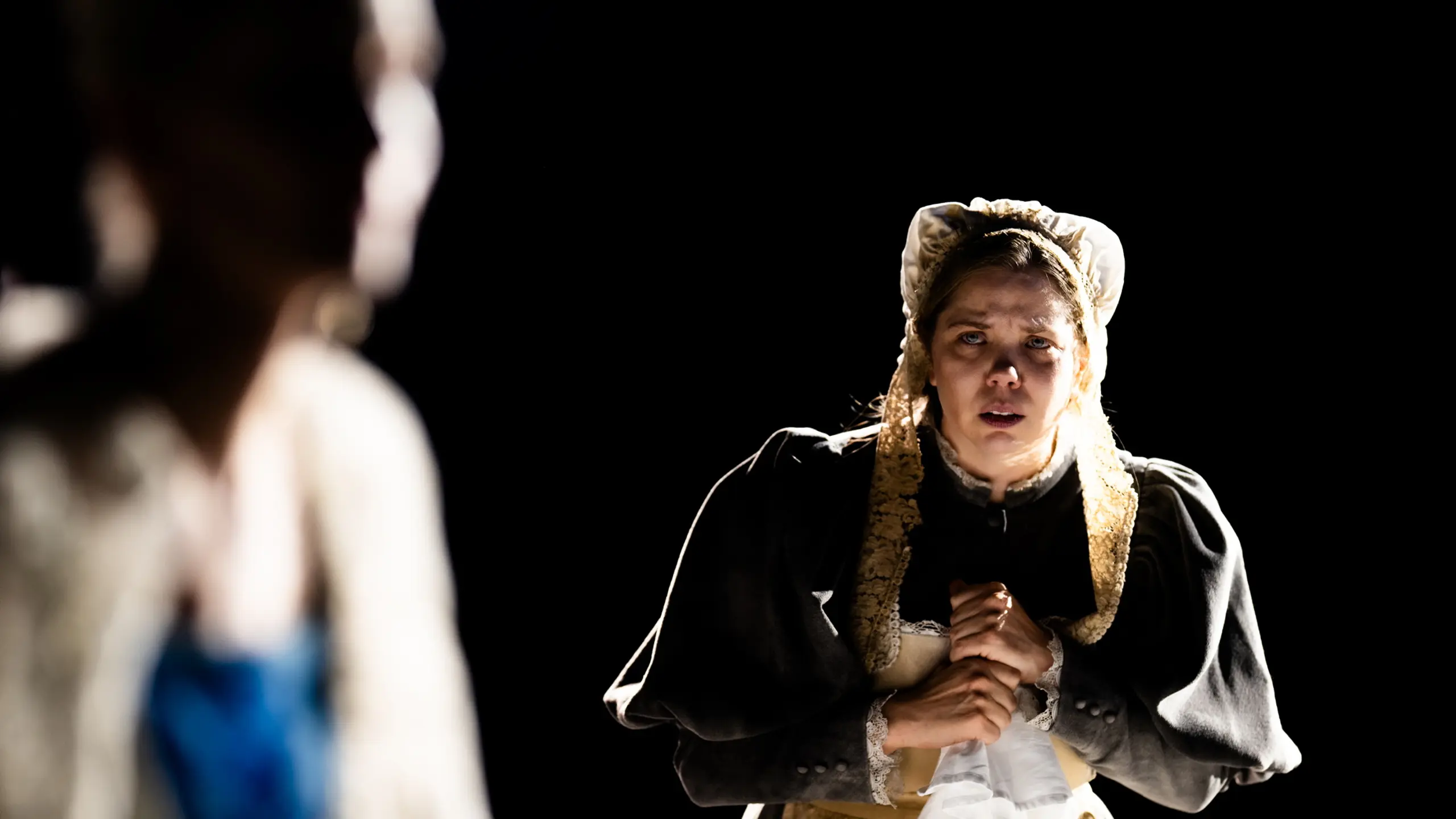 Kvinne i gammeldags kjole og kyse står med et hvitt lommetørkle og ser bekymret ut.