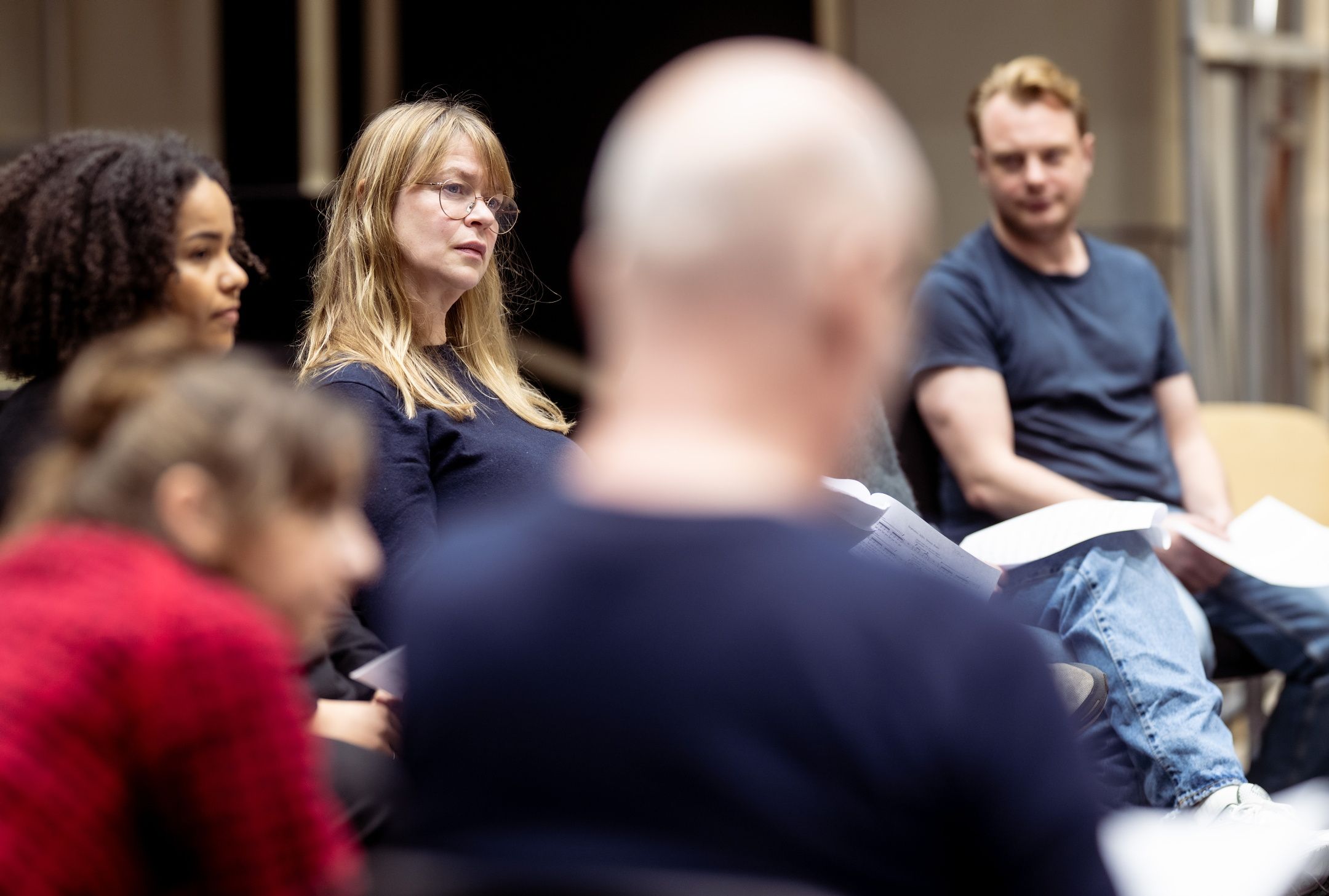 Flere skuespillere sitter i ring med manus i hånden og lytter oppmerksomt under en leseprøve.