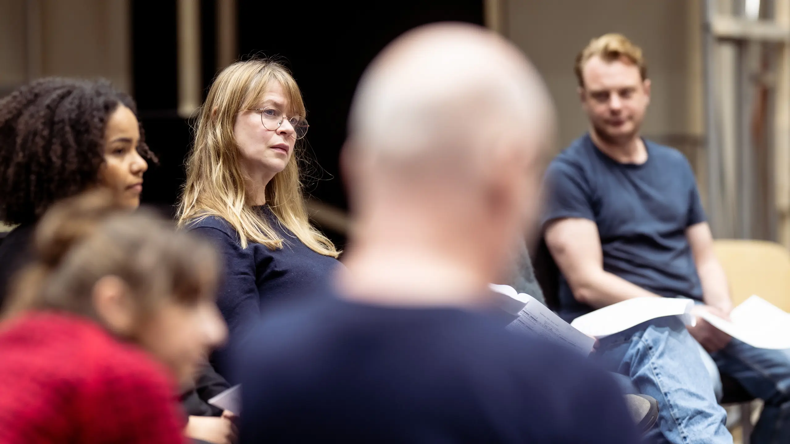 Flere skuespillere sitter i ring med manus i hånden og lytter oppmerksomt under en leseprøve.