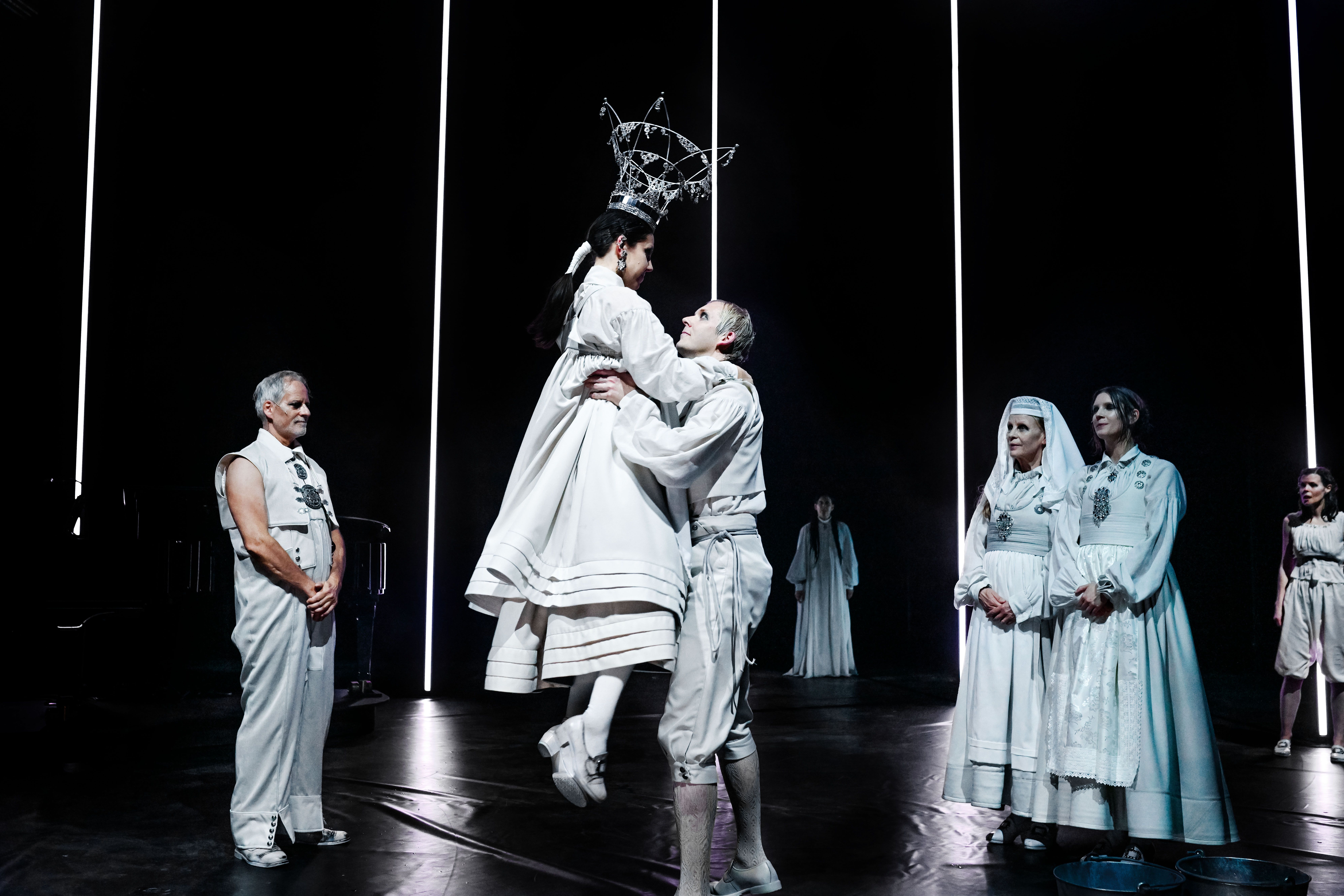 Syv sceneaktører i hvite bunadsinspirerte kostymer. Den ene mannen løfter opp en kvinne. Hun har brudekrone på