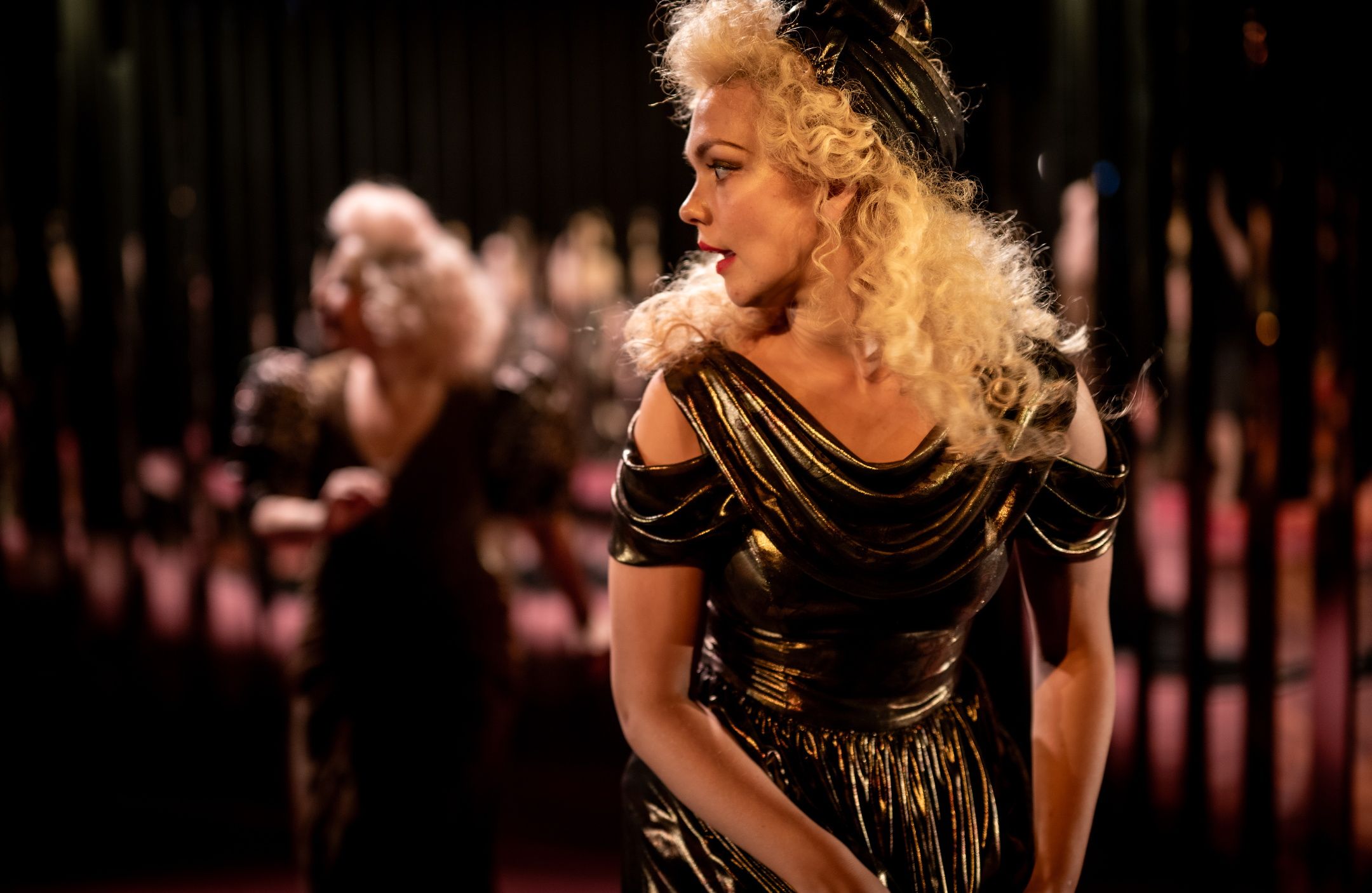 Kvinne i glinsende svart kjole og hårbånd ser til siden, mens en annen i samme antrekk er uskarp i bakgrunnen.