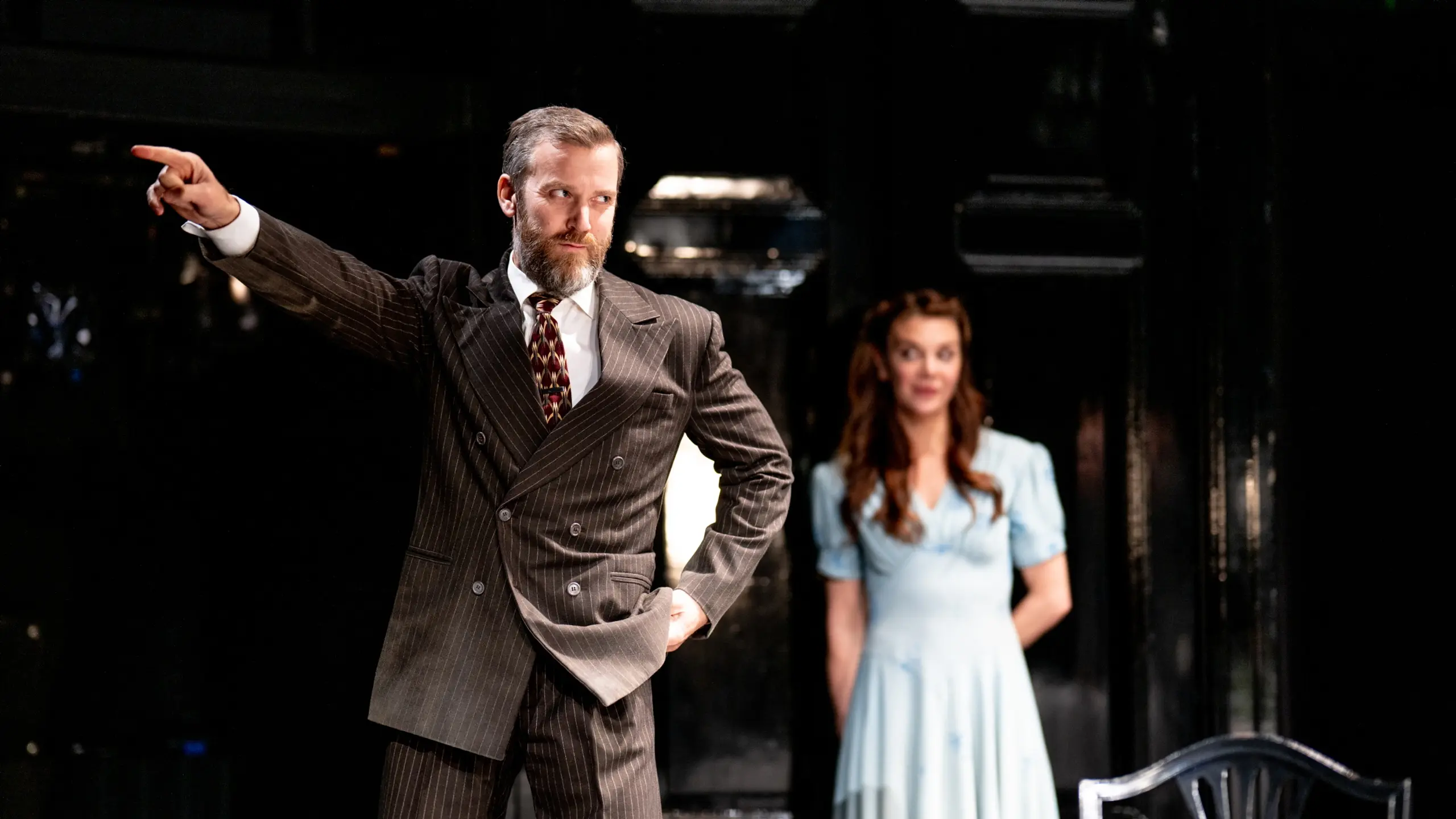 Mann i brun dress som peker. Kvinne i blå kjole i bakgrunnen