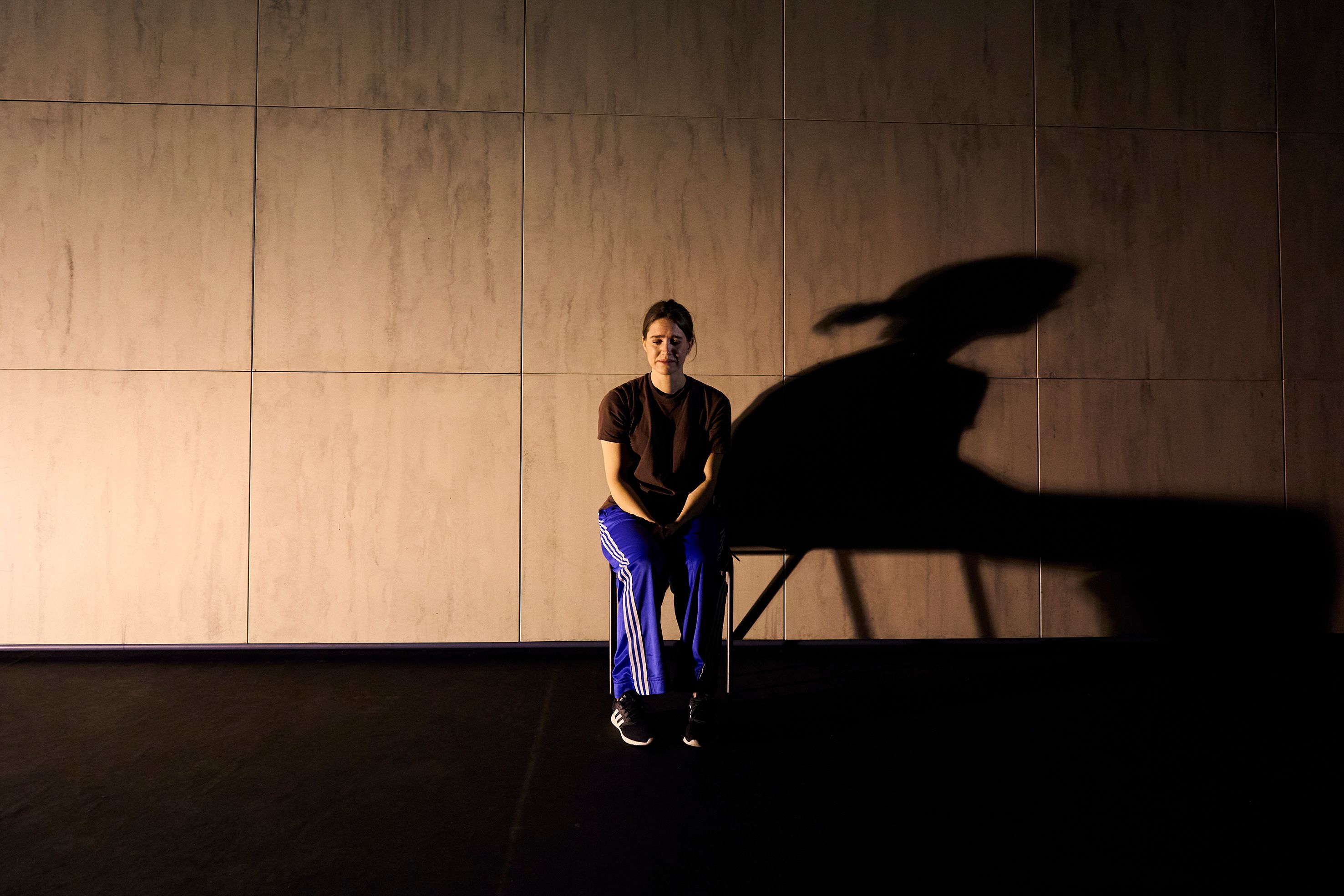 En kvinne sitter på en stol midt på scenen, med en stor skygge projisert på veggen bak henne.