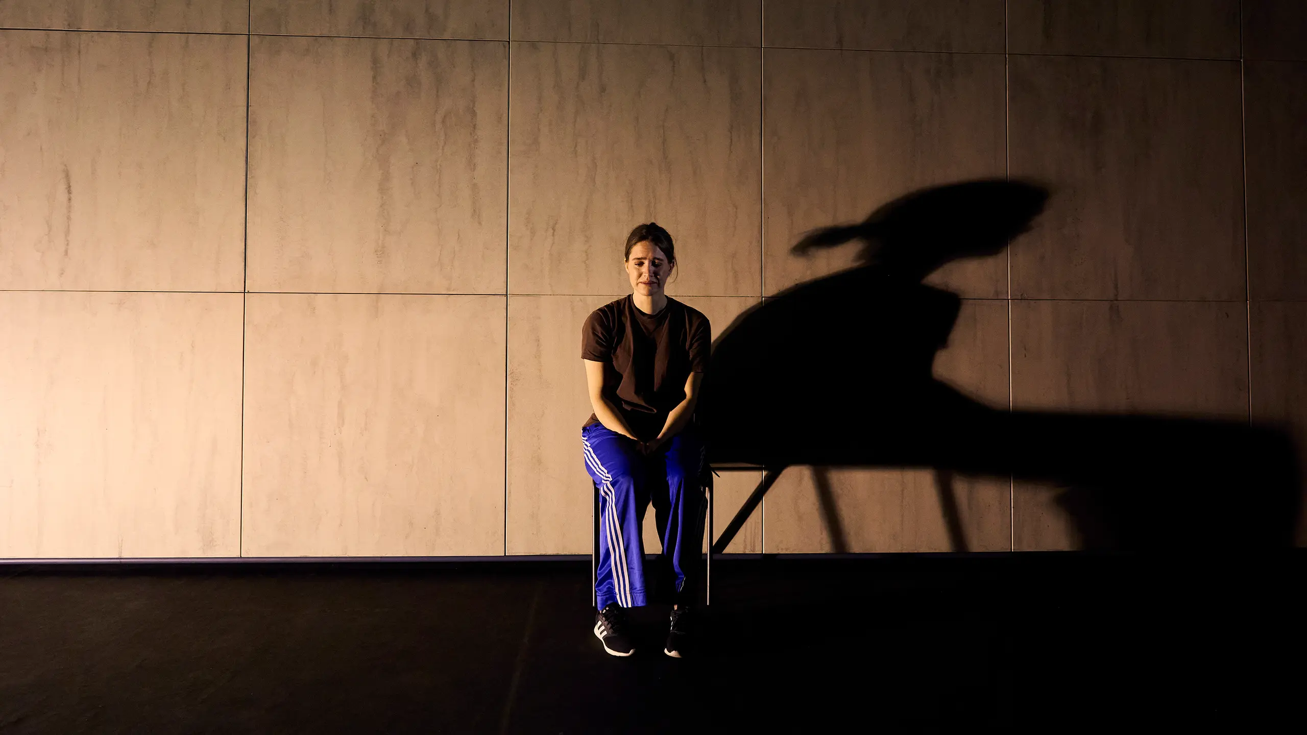 En kvinne sitter på en stol midt på scenen, med en stor skygge projisert på veggen bak henne.