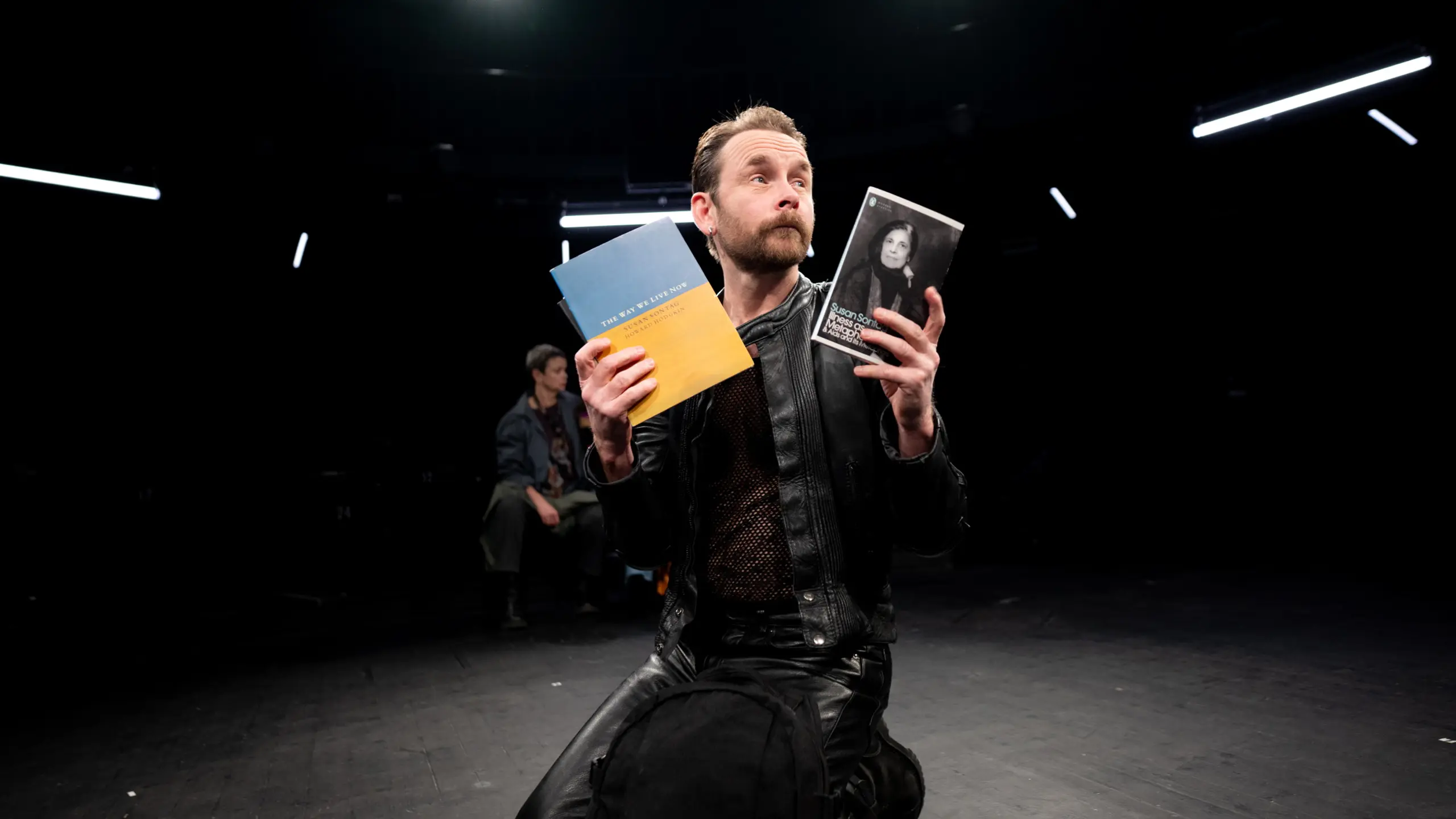 Mann i skinnjakke kneler på scenen og holder opp to bøker med alvorlig ansiktsuttrykk.