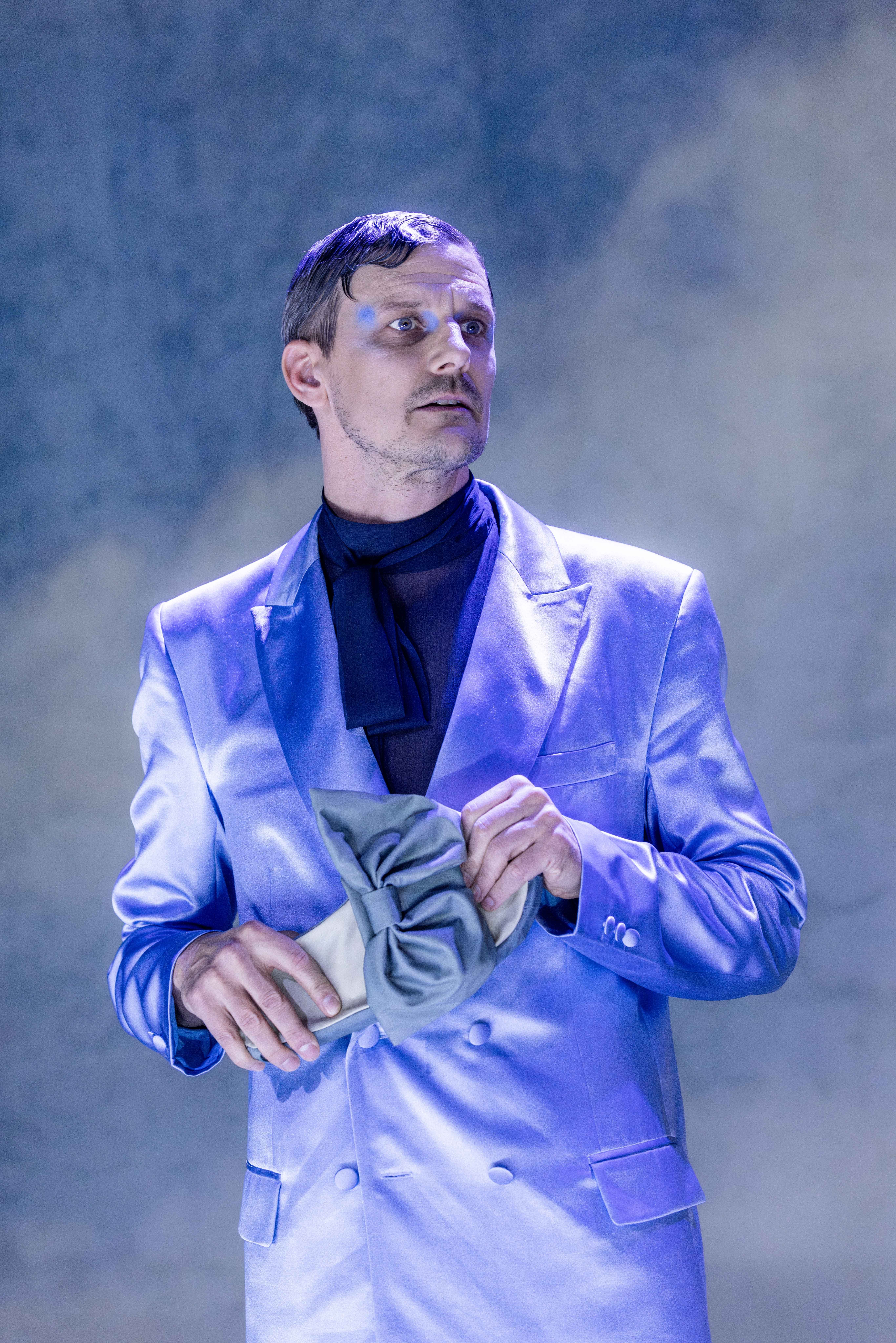 Person i lys blå dress holder en liten veske med stor sløyfe, ser opp mot siden i scenelys