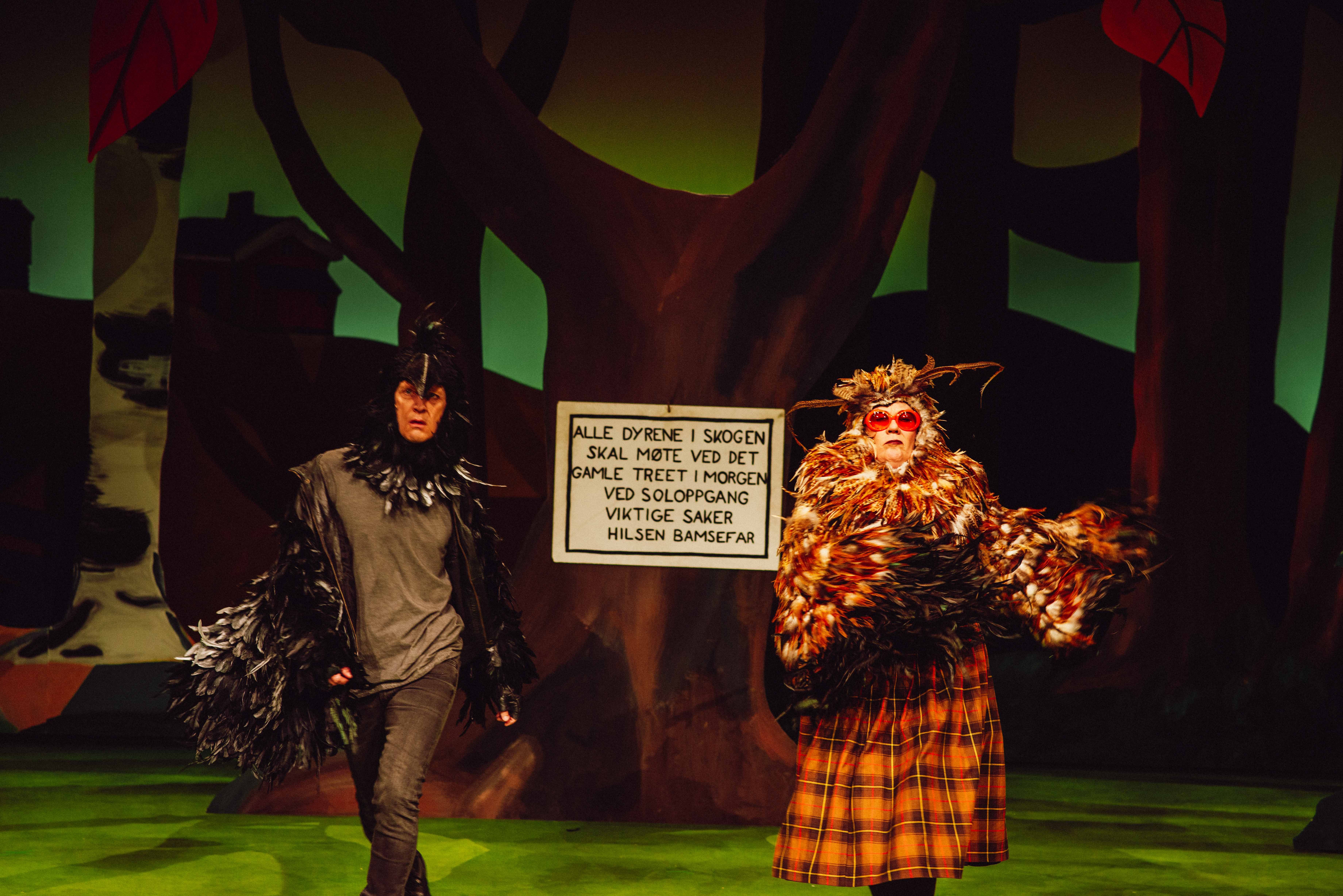 To personer i fuglekostymer foran skilt med teksten 'Alle dyrene i skogen... Hilsen Bamsefar'.