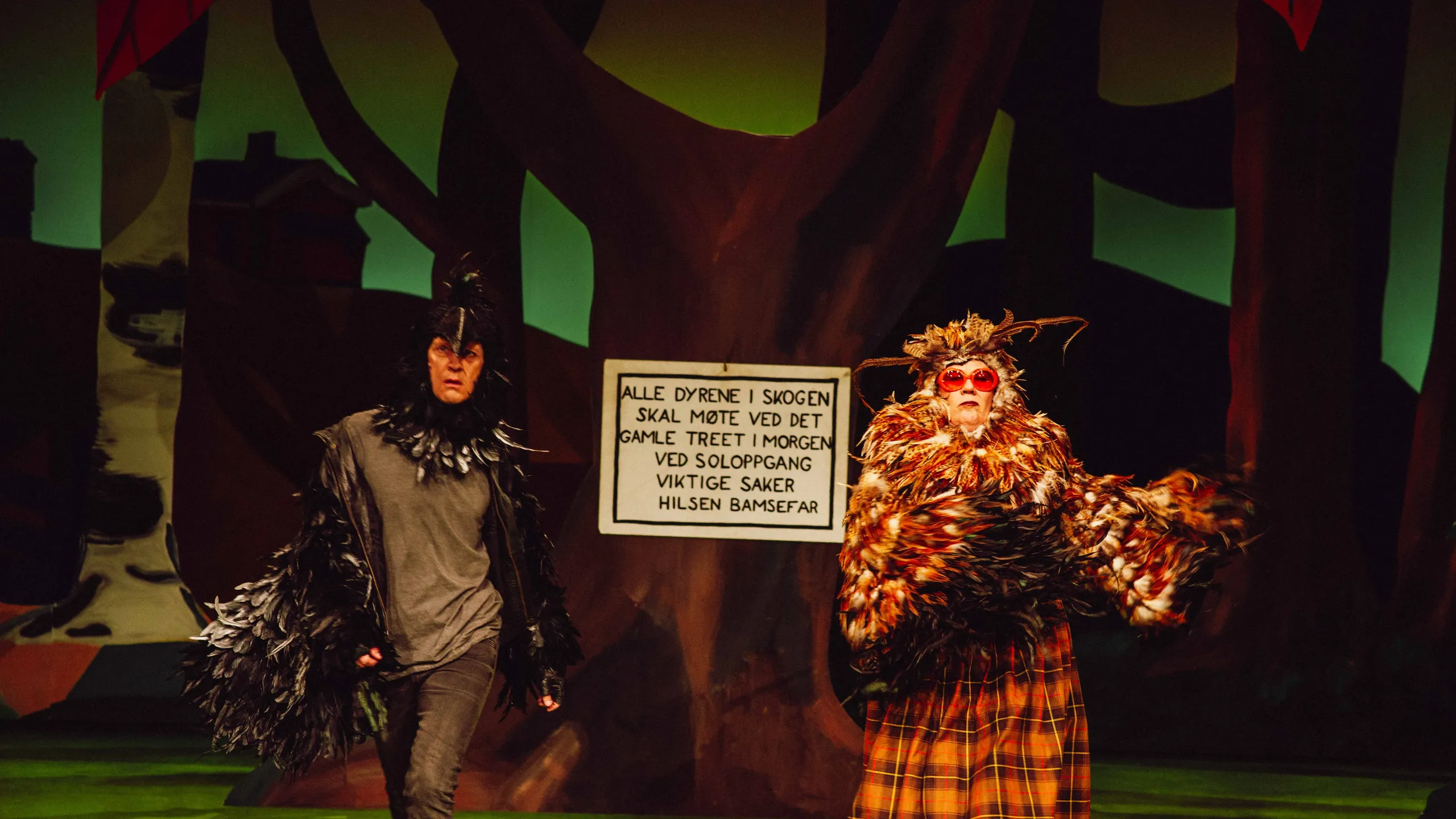 To personer i fuglekostymer foran skilt med teksten 'Alle dyrene i skogen... Hilsen Bamsefar'.