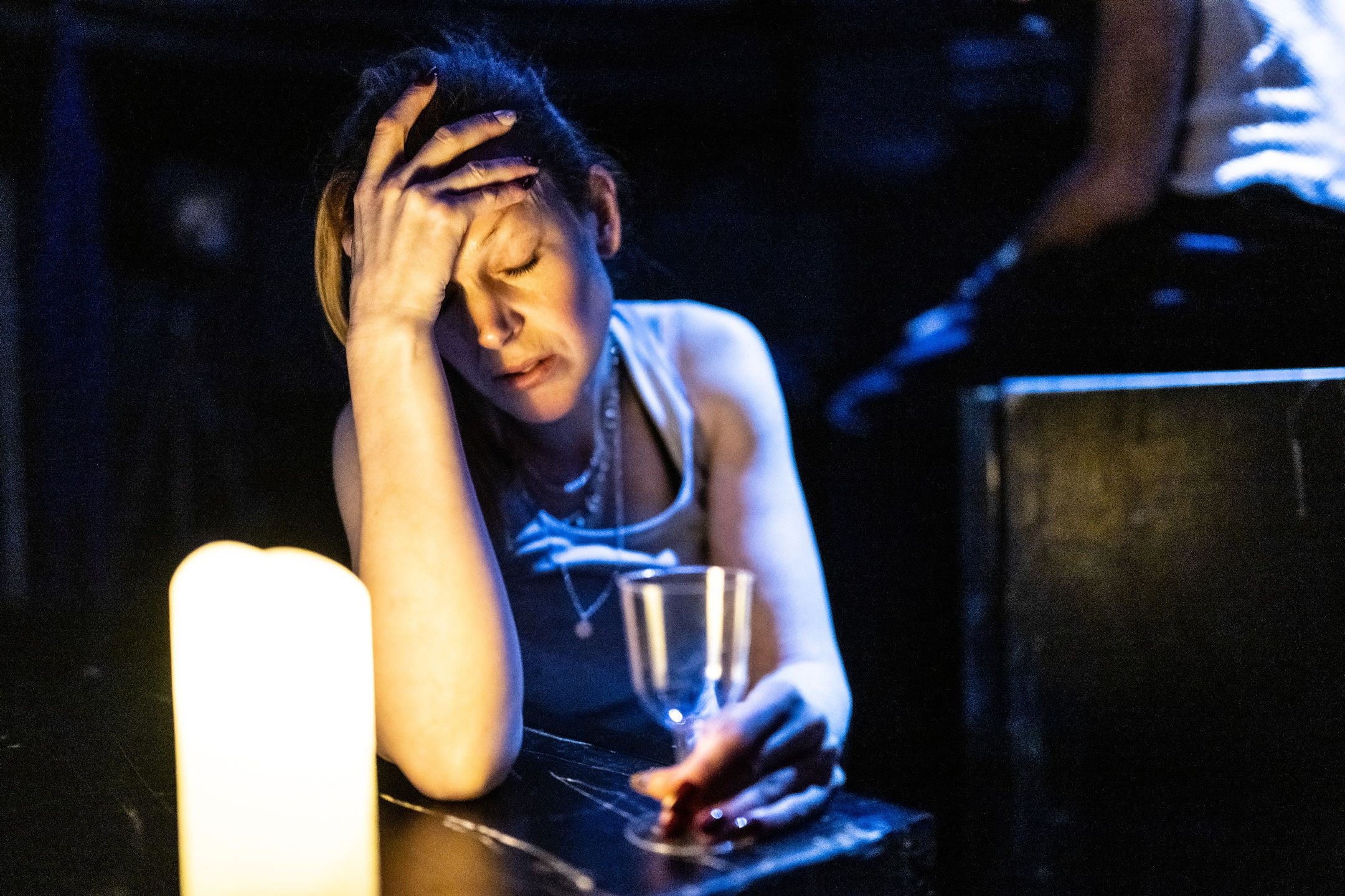 Kvinne med glass i hånden sitter ved tente lys og holder hodet i hånden med lukket øyne