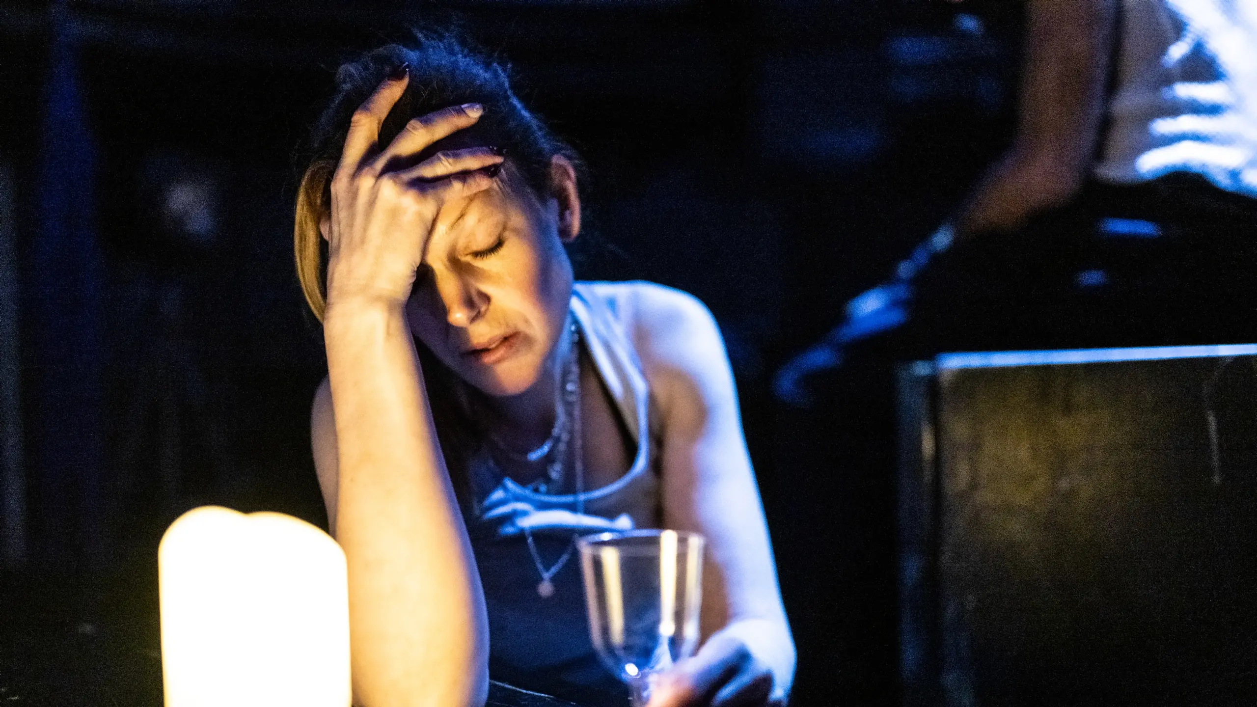 Kvinne med glass i hånden sitter ved tente lys og holder hodet i hånden med lukket øyne