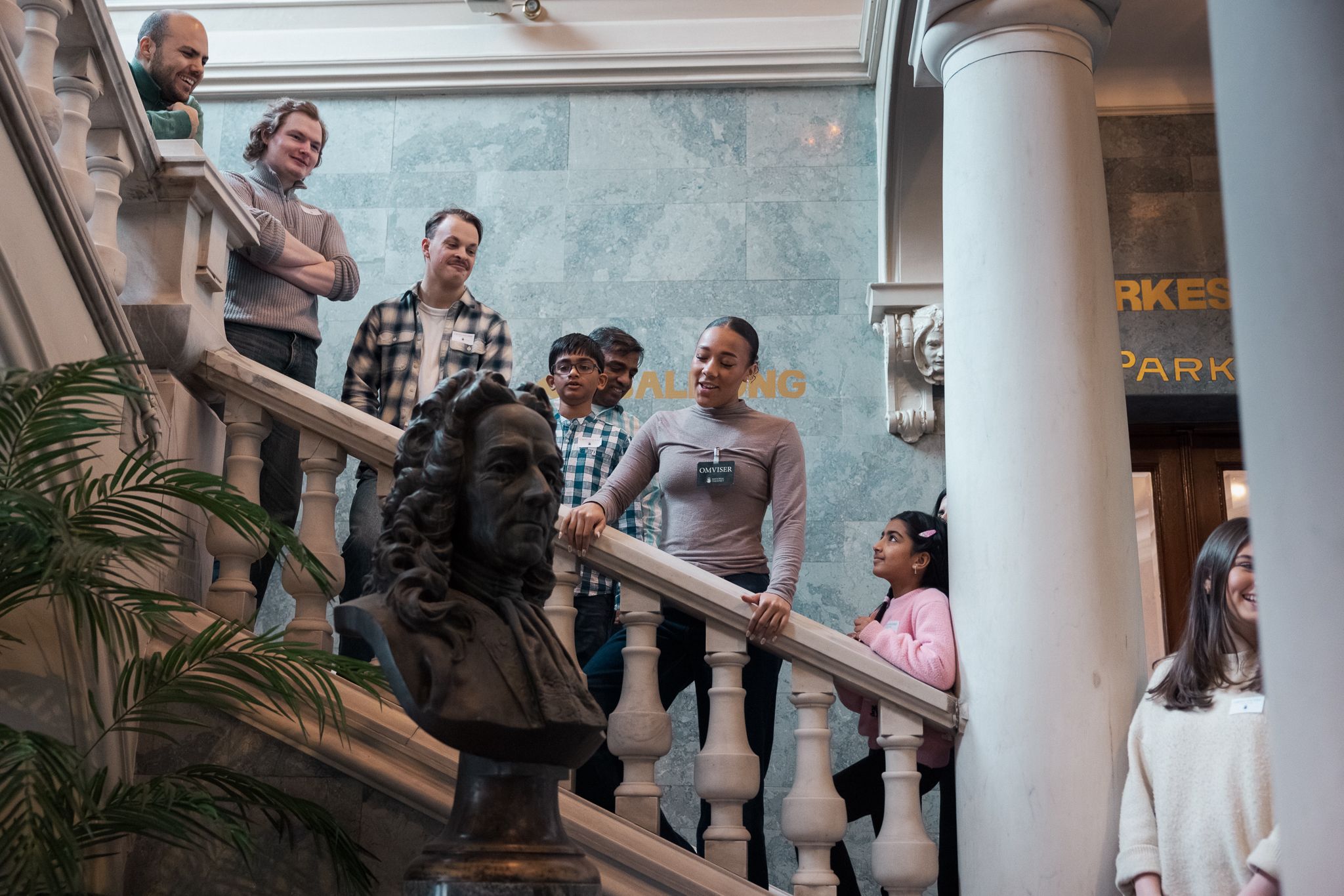 Gruppe med barn og voksne står i trapp med marmorrekkverk og byste i forgrunnen, omviser forklarer.
