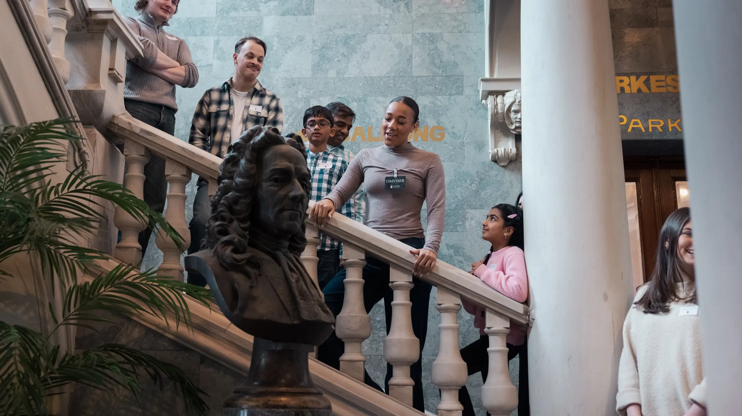 Gruppe med barn og voksne står i trapp med marmorrekkverk og byste i forgrunnen, omviser forklarer.