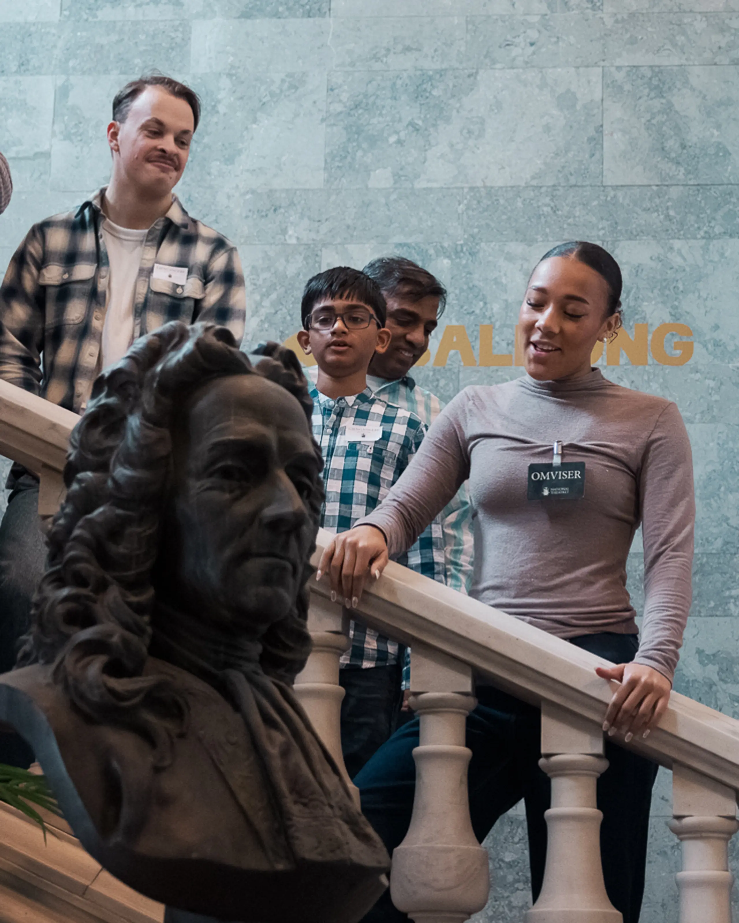 Gruppe med barn og voksne står i trapp med marmorrekkverk og byste i forgrunnen, omviser forklarer.