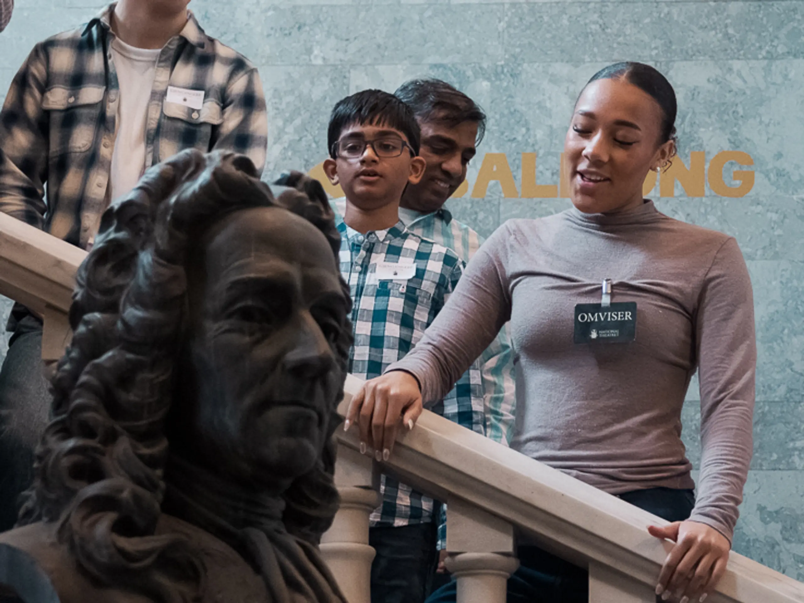 Gruppe med barn og voksne står i trapp med marmorrekkverk og byste i forgrunnen, omviser forklarer.