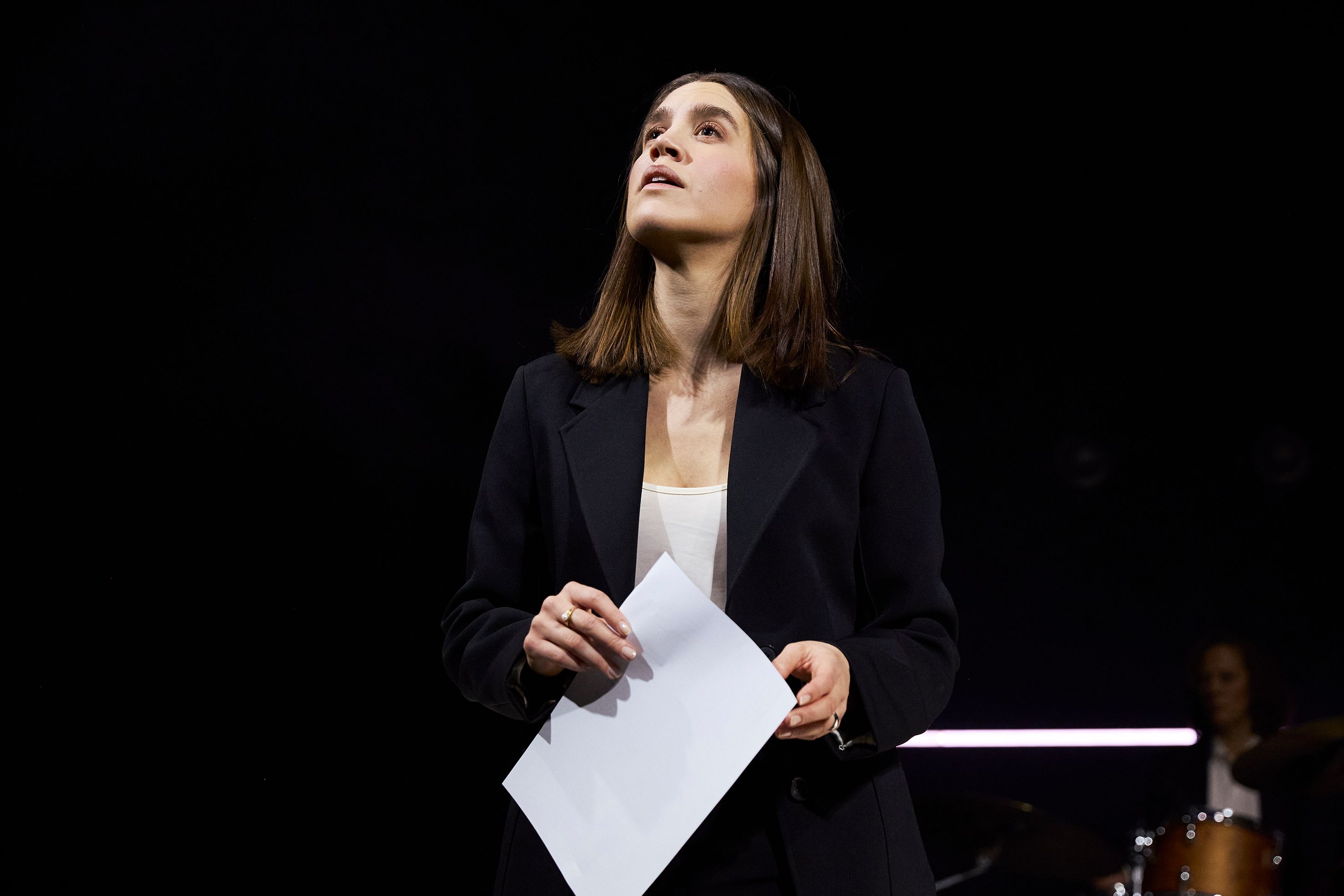 En kvinne i svart dress holder et ark og ser oppover med et tankefullt uttrykk.