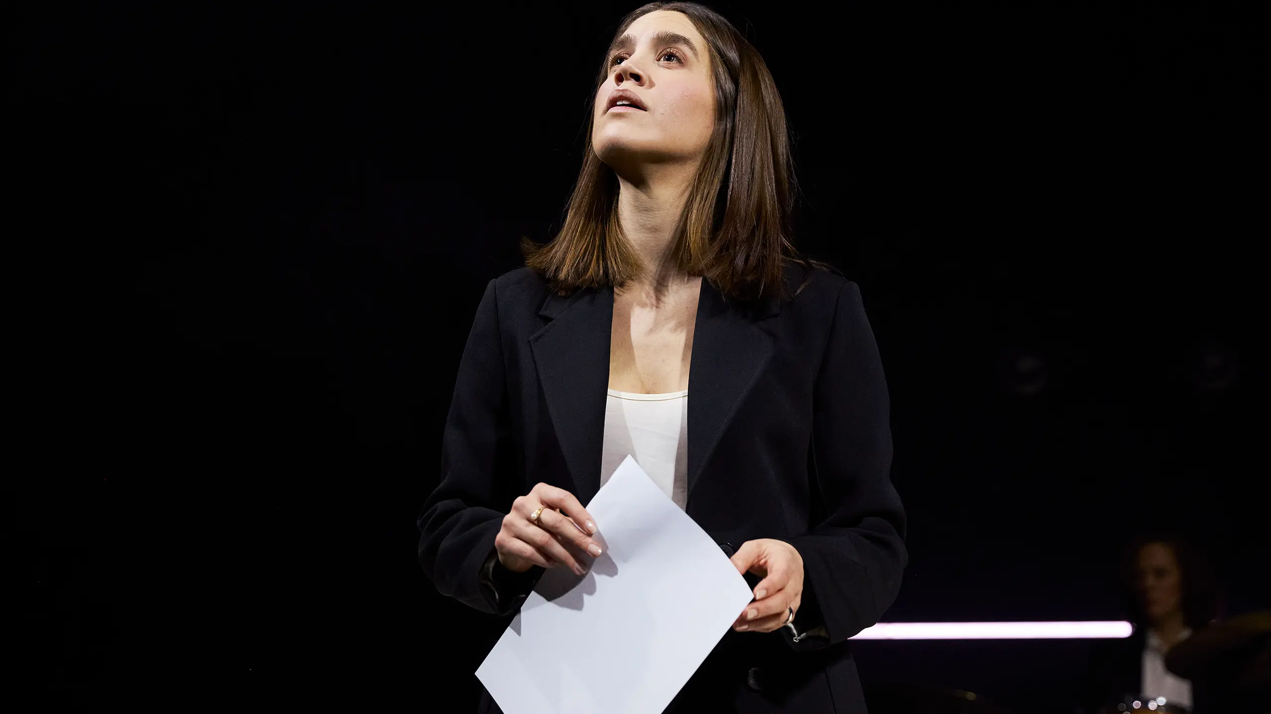 En kvinne i svart dress holder et ark og ser oppover med et tankefullt uttrykk.