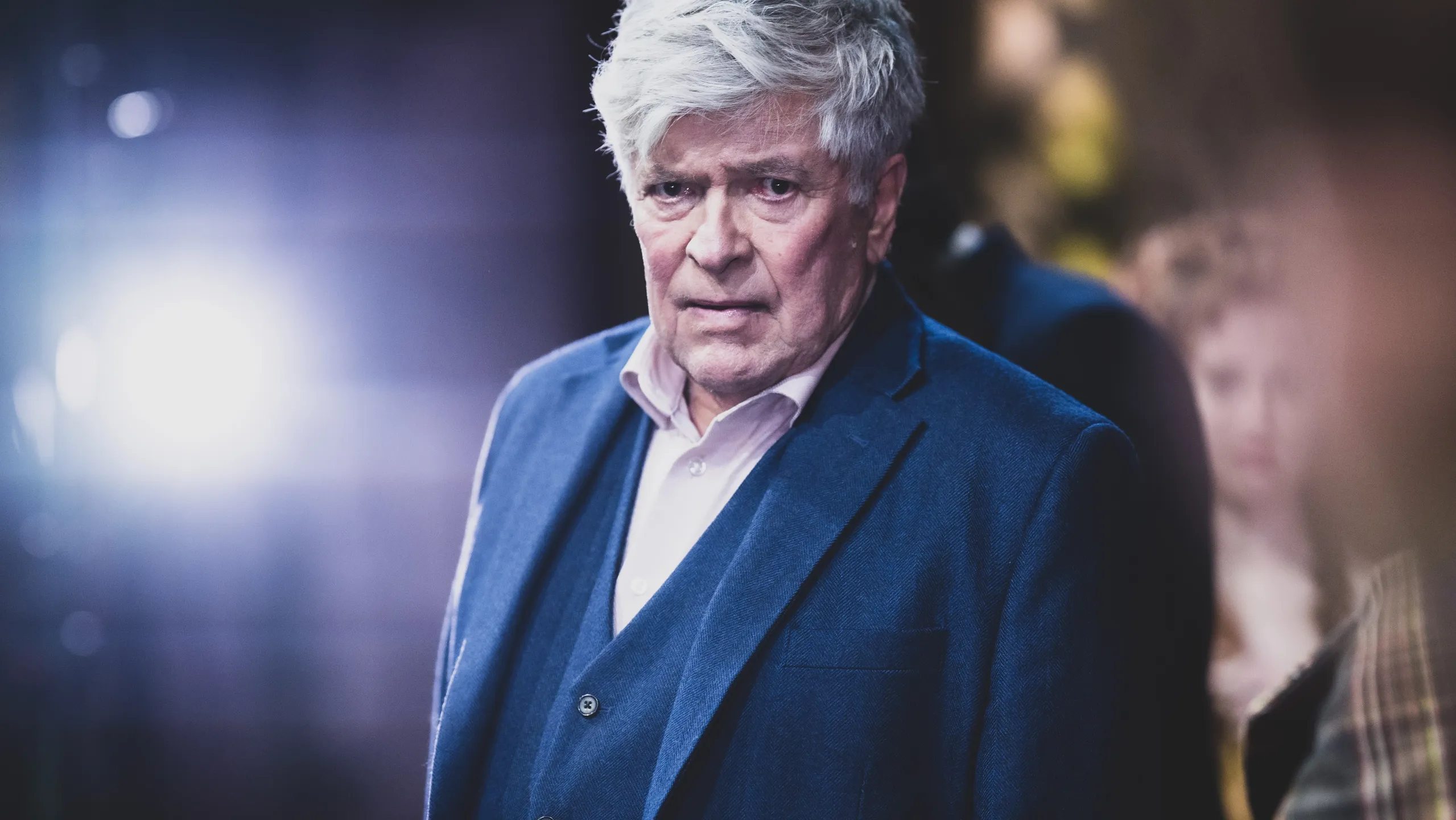 person med kort grått hår i blå dress ser alvorlig fremover i scenelys