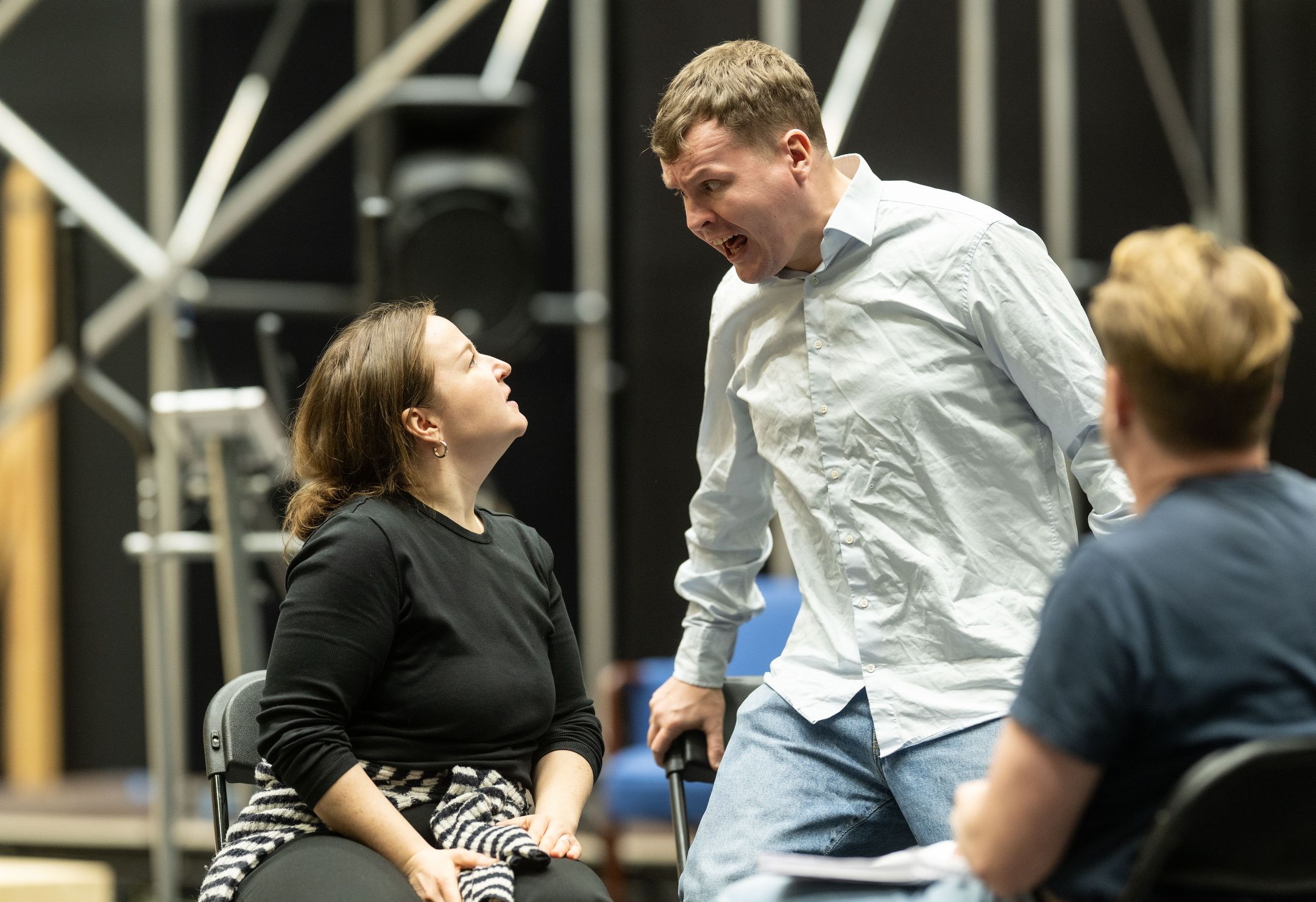 En kvinne i svart og en mann i lys skjorte fremfører en konfronterende scene, mens en tredje person ser på.