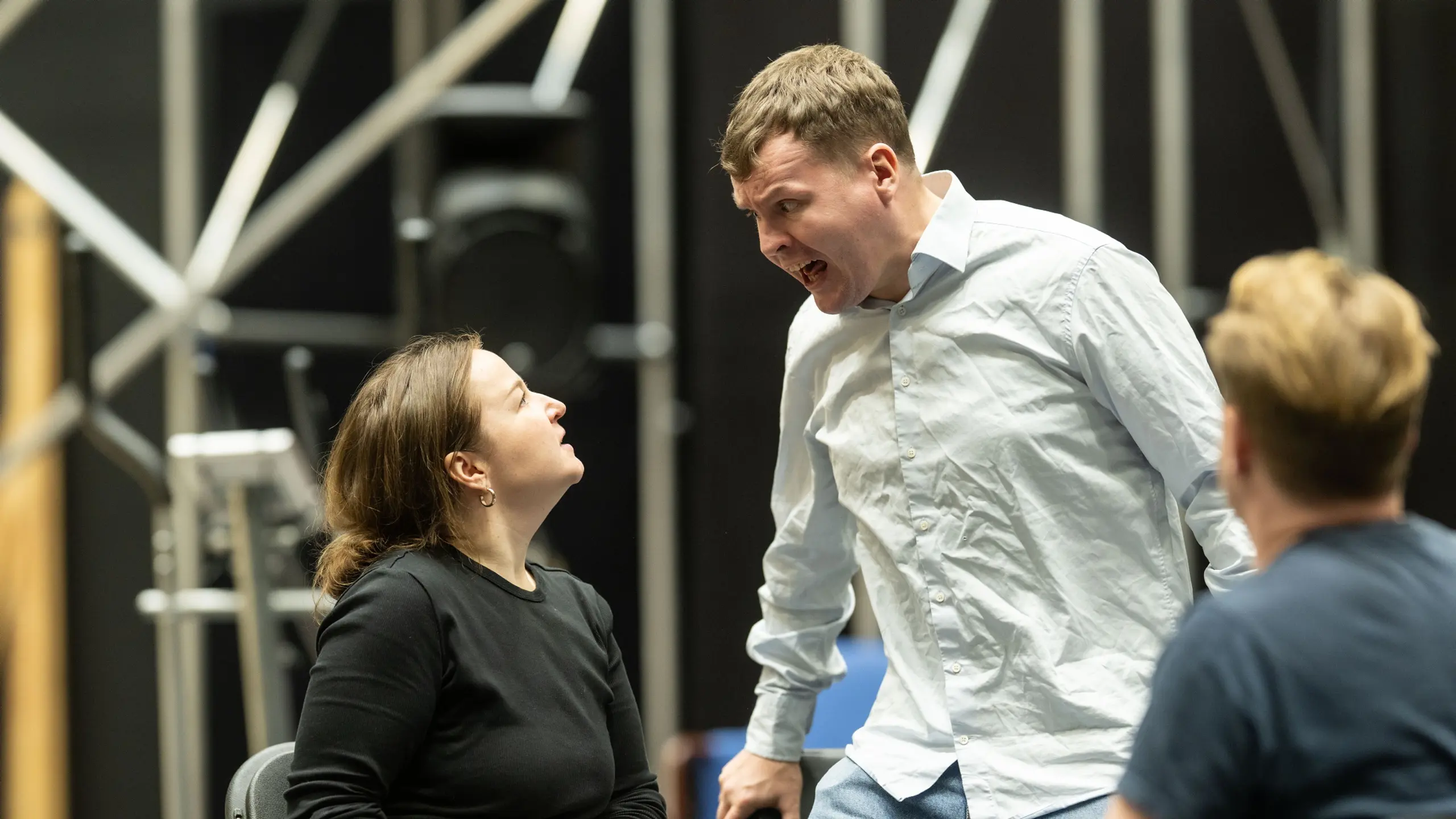 En kvinne i svart og en mann i lys skjorte fremfører en konfronterende scene, mens en tredje person ser på.