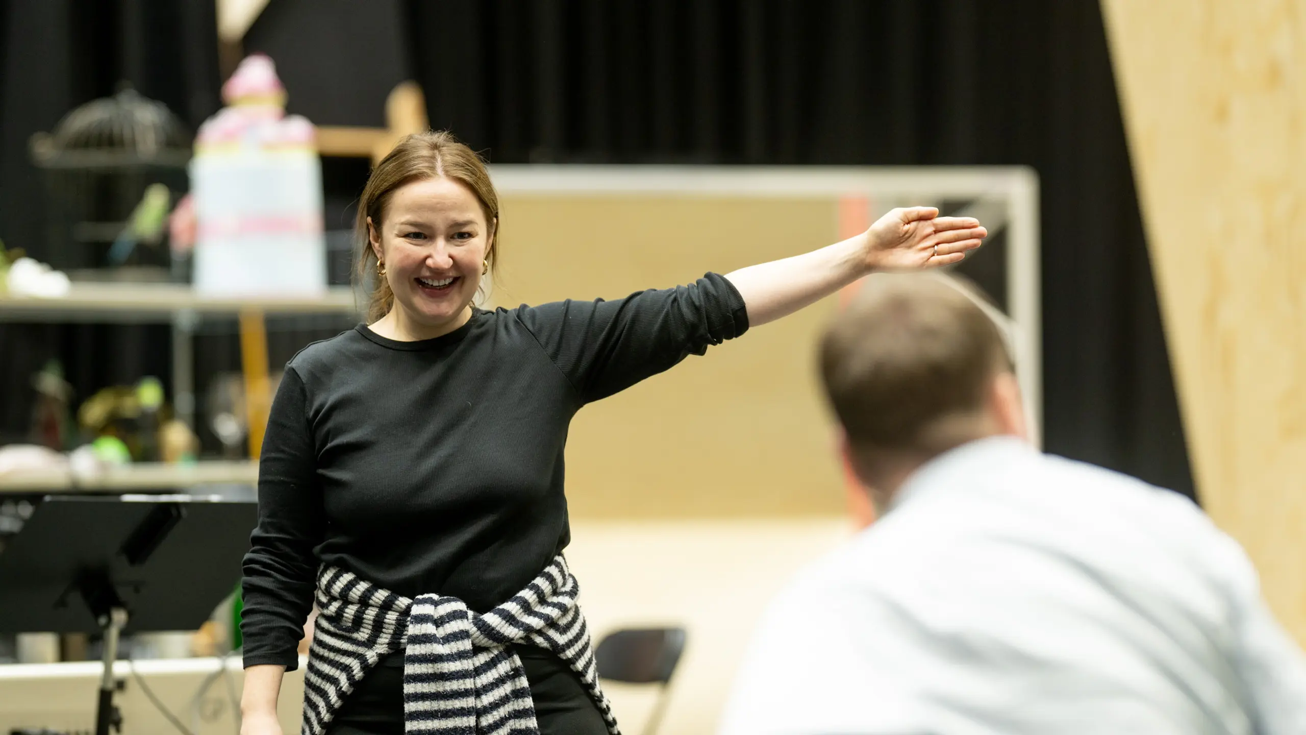 En kvinne i svart genser smiler og gestikulerer mot en mann i lyseblå skjorte med ryggen til kamera.