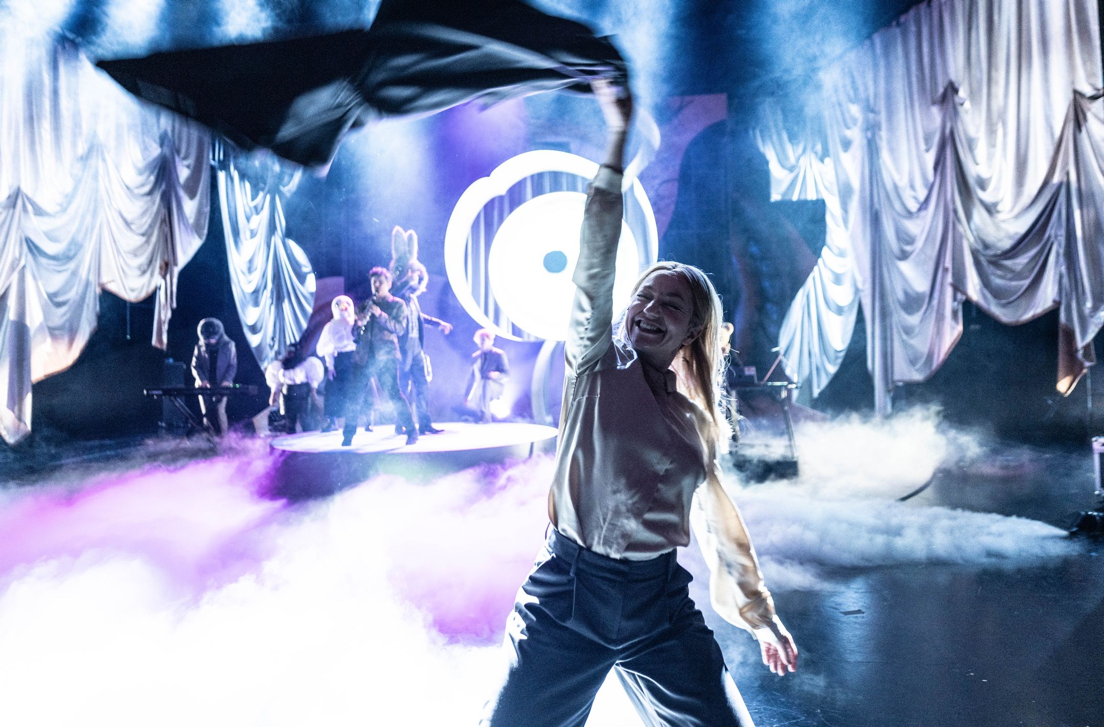 Person smiler og veiver med jakke i forgrunnen, mens flere karakterer opptrer i tåkelagt, lystett scenebilde bak.