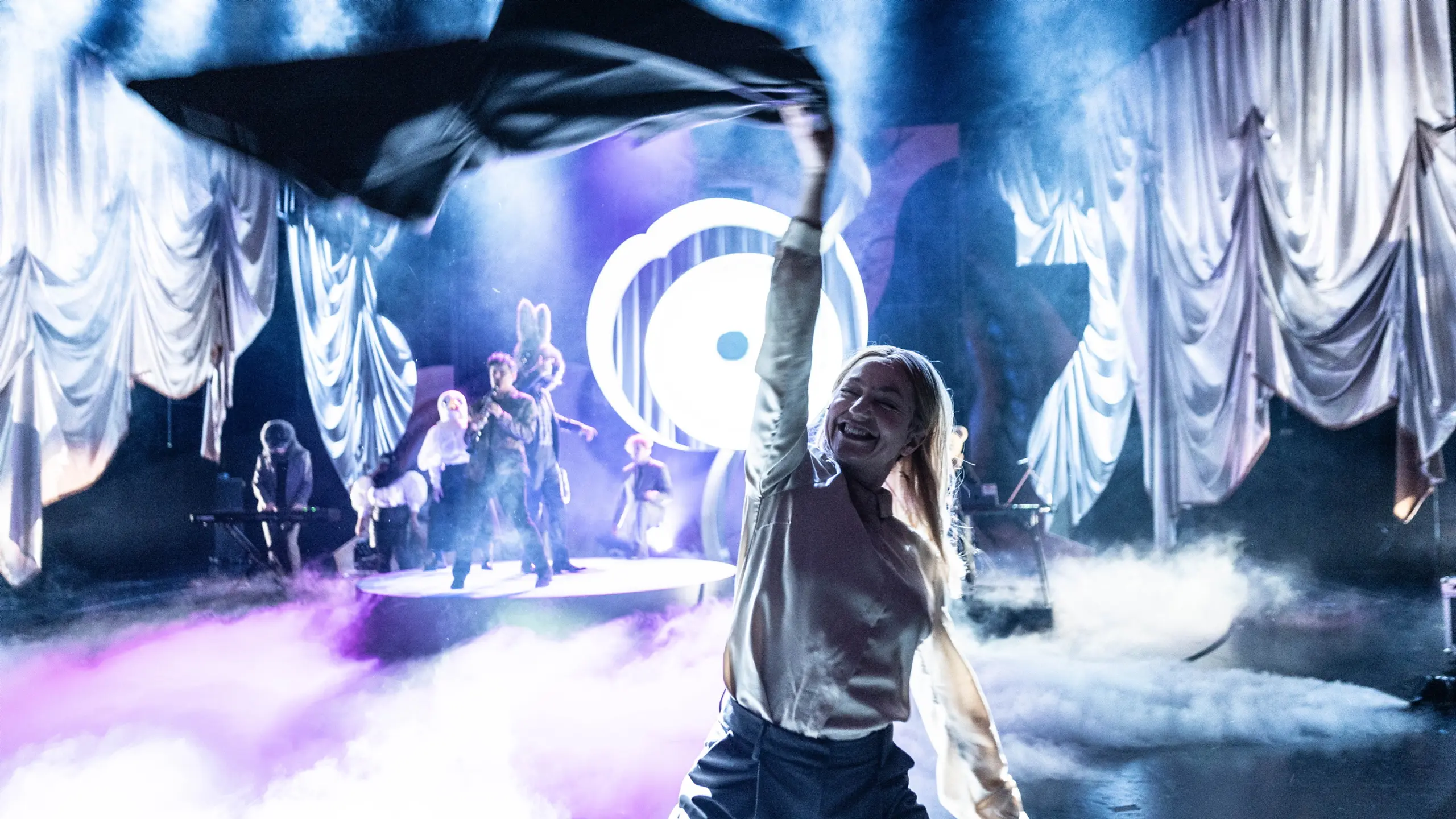 Person smiler og veiver med jakke i forgrunnen, mens flere karakterer opptrer i tåkelagt, lystett scenebilde bak.