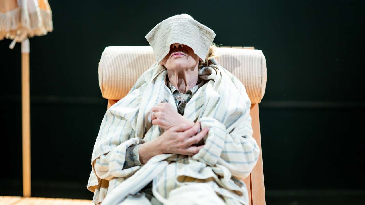 kvinne med håndkle over øynene sitter tilbakelent i solseng på scenegulv.