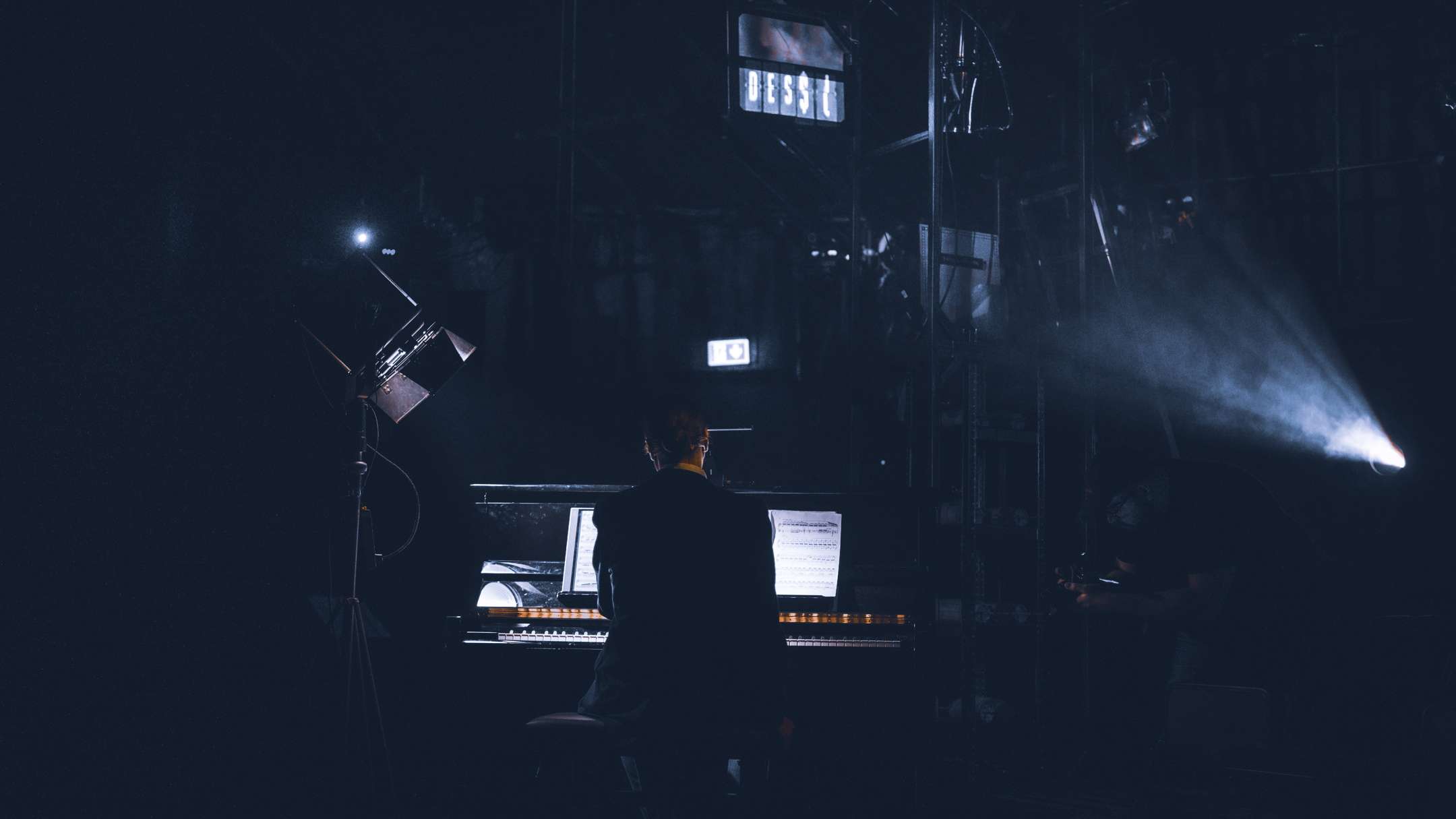 Mann sitter ved piano i scenelys med ryggen til kamera