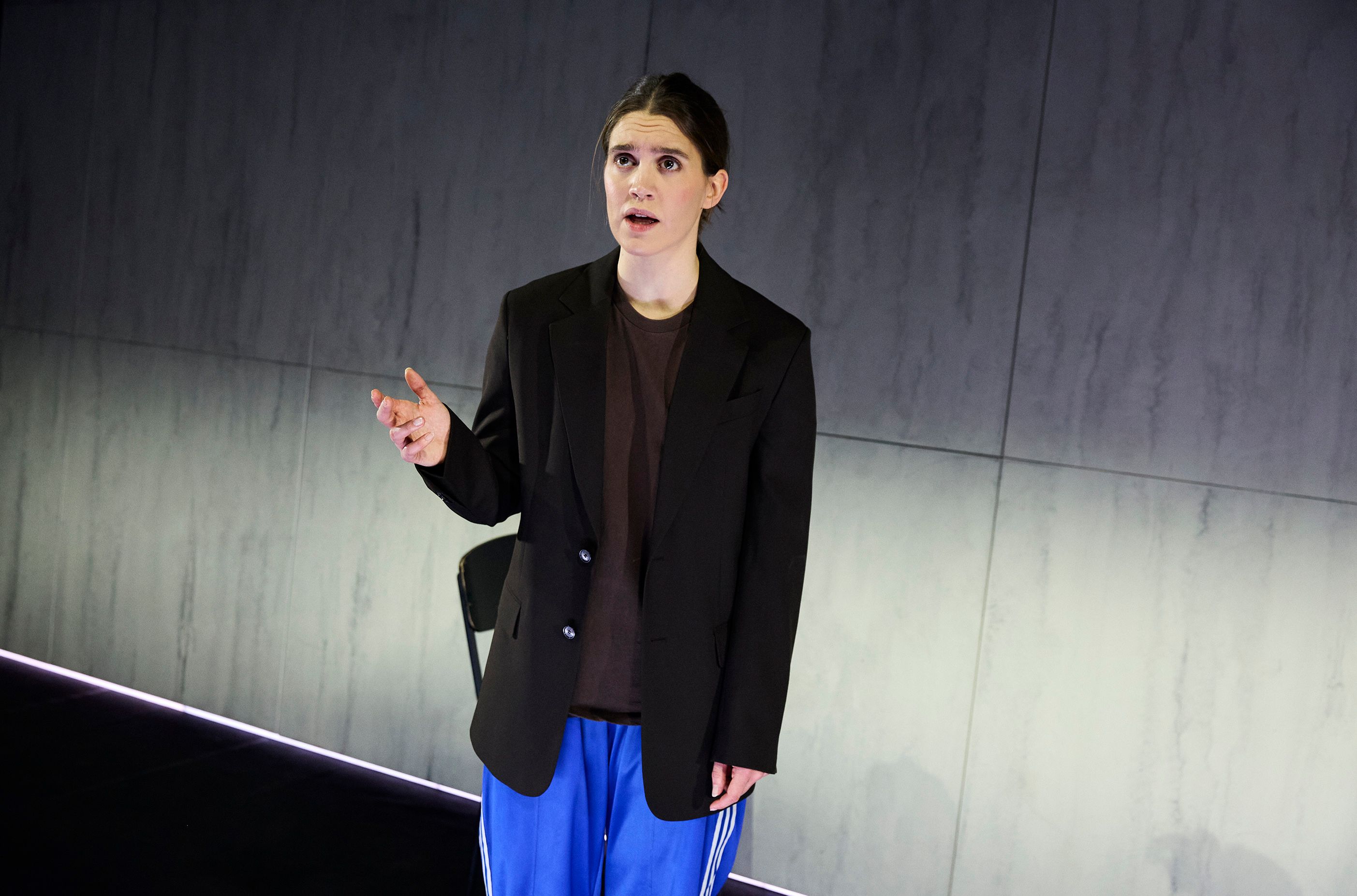 En kvinne med sort blazer, brun t-skjorte og blå treningsbukser gestikulerer med den ene hånden mens hun snakker på scenen.