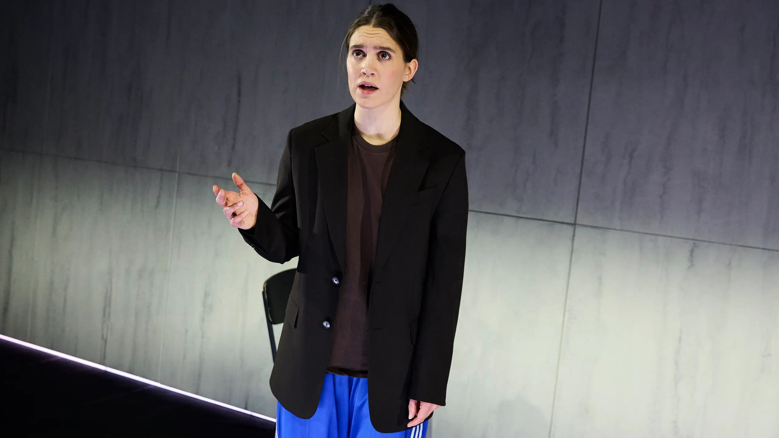 En kvinne med sort blazer, brun t-skjorte og blå treningsbukser gestikulerer med den ene hånden mens hun snakker på scenen.