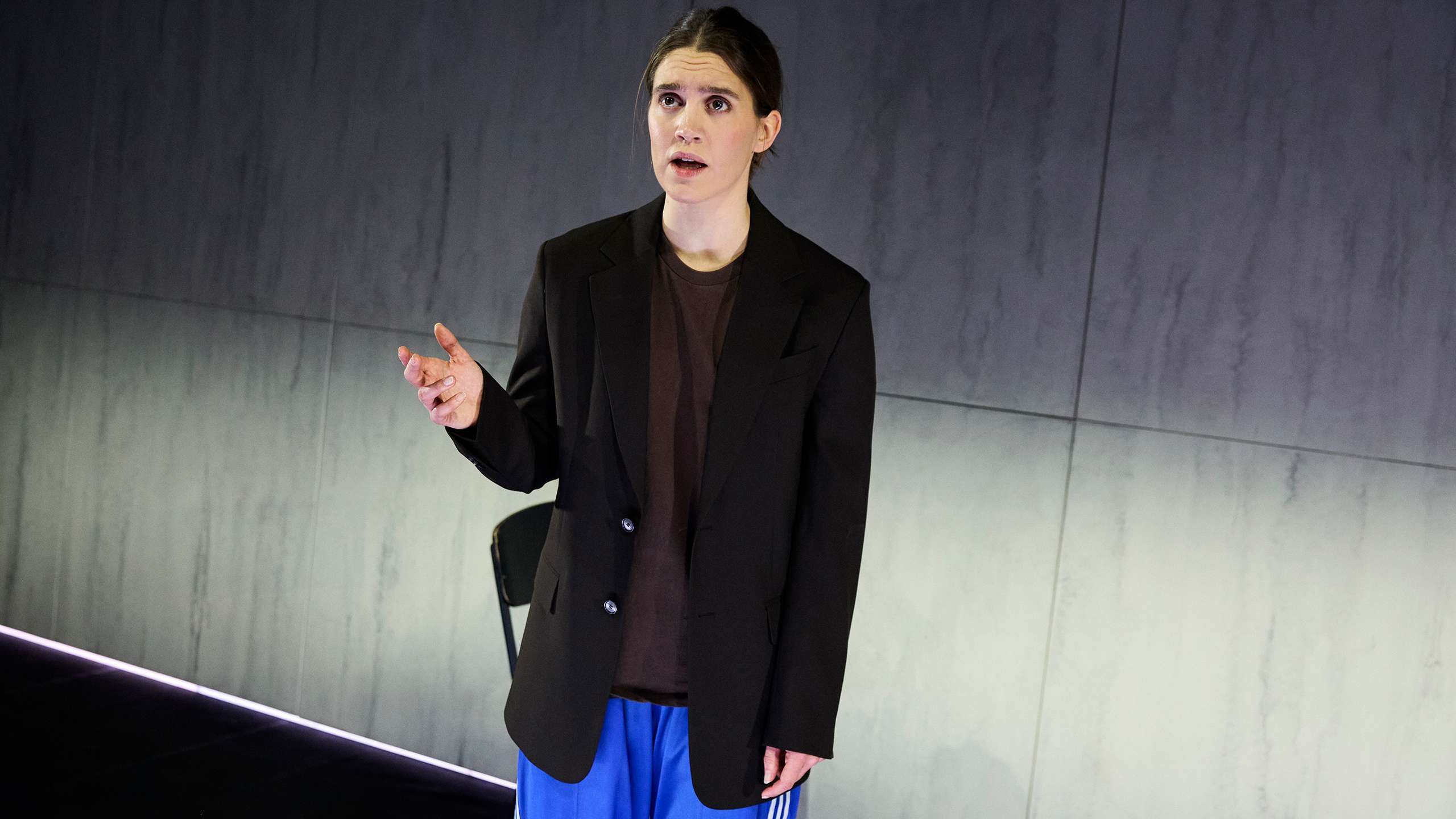 En kvinne med sort blazer, brun t-skjorte og blå treningsbukser gestikulerer med den ene hånden mens hun snakker på scenen.