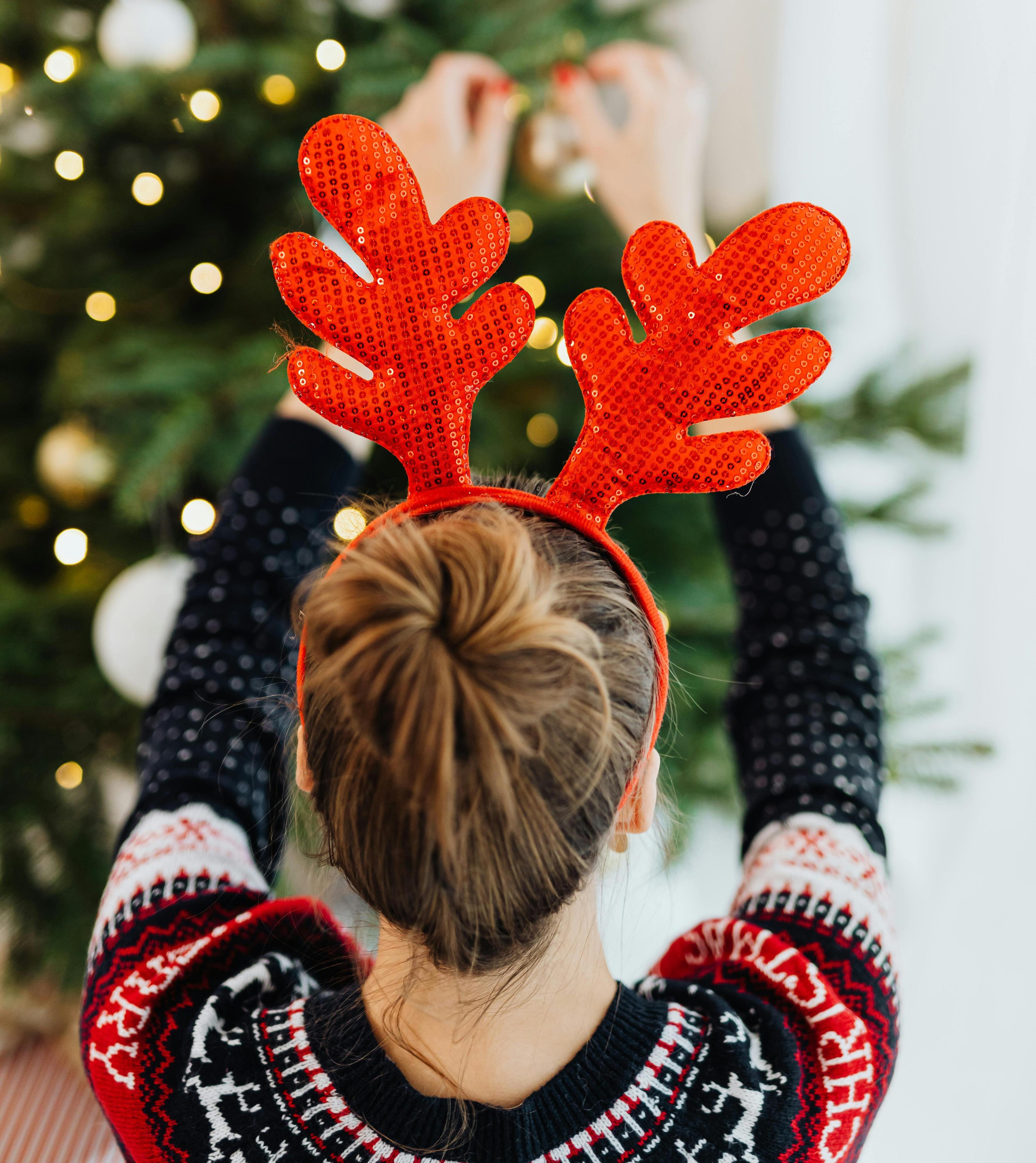 rénszarvasdísz egy nő fején, aki egy karácsonyfát díszít