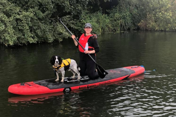 Nicola is kneeling on a bright red paddleboard, wearing a matching red life jacket, with Sprocker, Bob, wearing a yellow doggy life jacket, standing on the paddleboard in front of her. The water is calm and behind them is a large covering of bushes and trees.