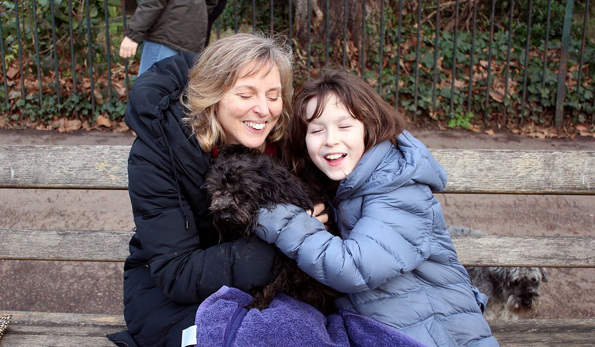 A woman and child are sitting on a park bench cuddling a black, curly-haired dog