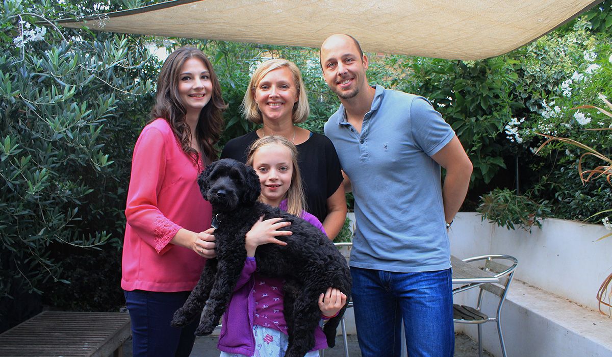 Three adults and a child are standing on a patio. The child is holding a medium sized, black, curly-haired dog.