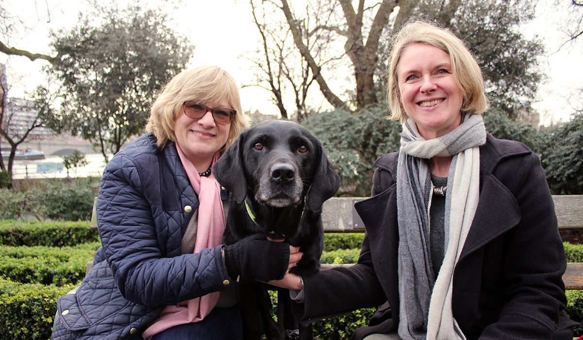 Two blonde women sitting on a bench, smiling, with a black dog between them