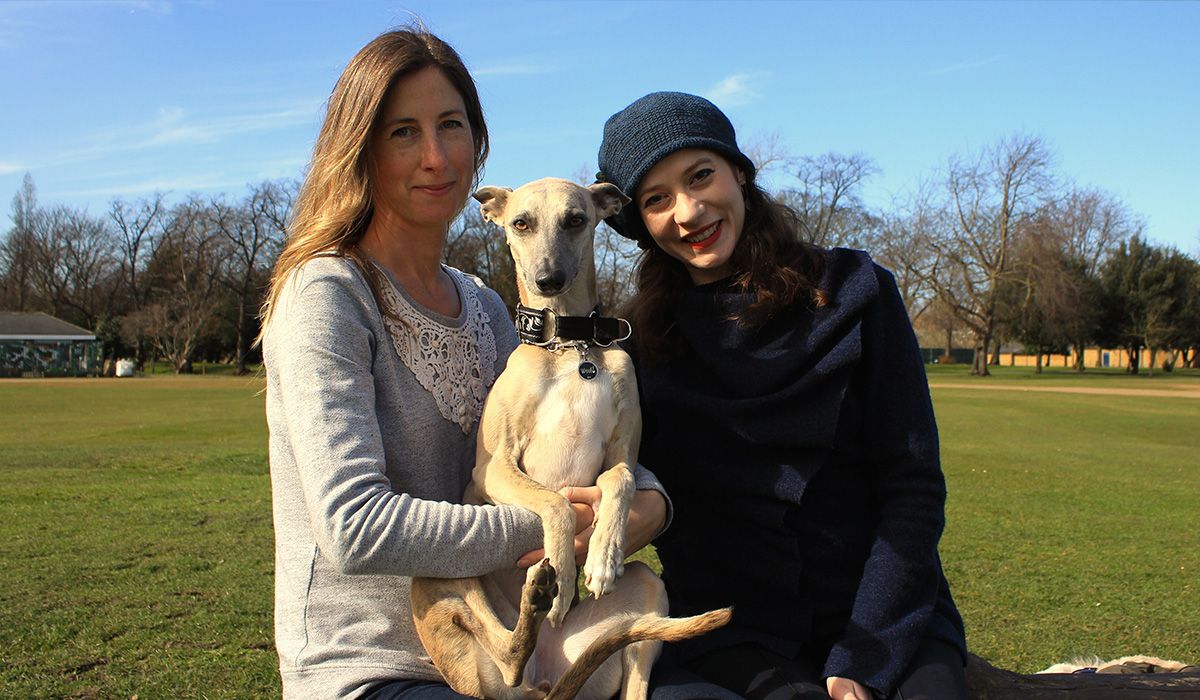 Two women are sitting in a park holding a slim dog who seems to be all legs!