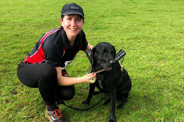 Runner, Nicola, is crouched beside black Labrador, Lola, holding a gold medal around Lola's chest proudly after winning their canicross race.