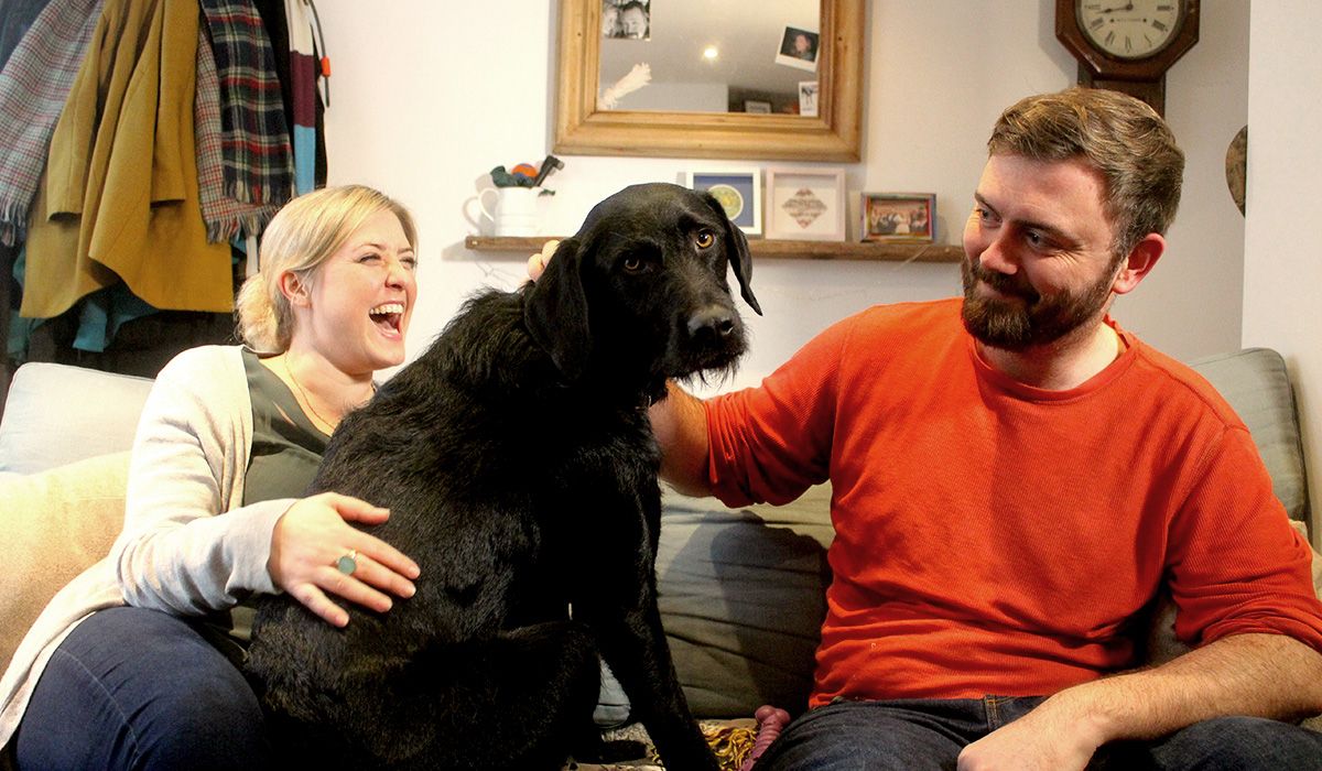 Two people and a dog are sitting on a sofa in a home.
