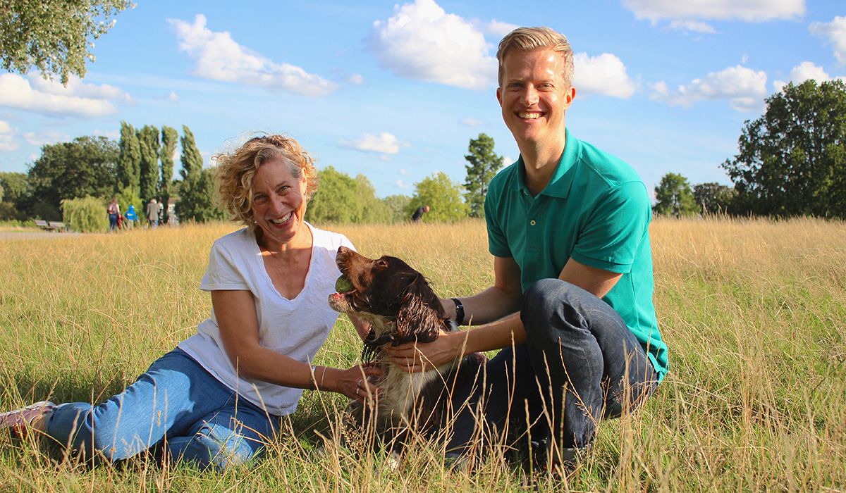 Two smiling people and a brown and white dog are sitting on grass in a park.