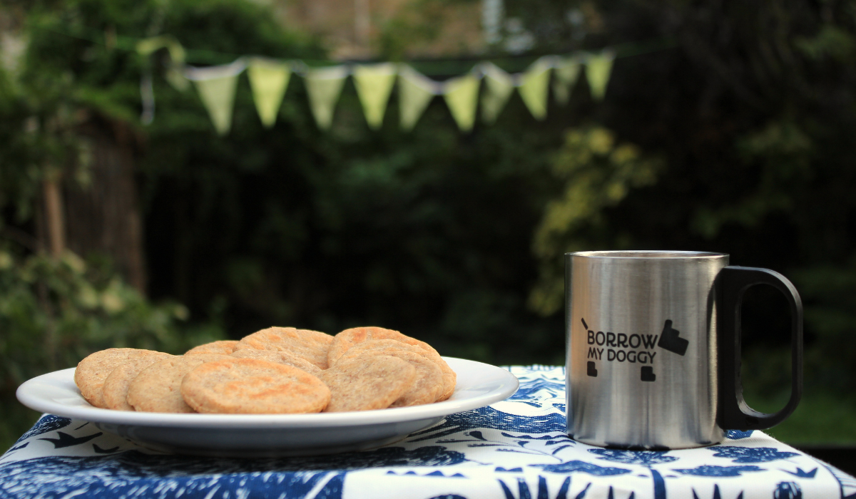 A plate of Peanut Butter Cookies next to a BorrowMyDoggy silver mug, on a table in the garden with bunting hanging in the background.