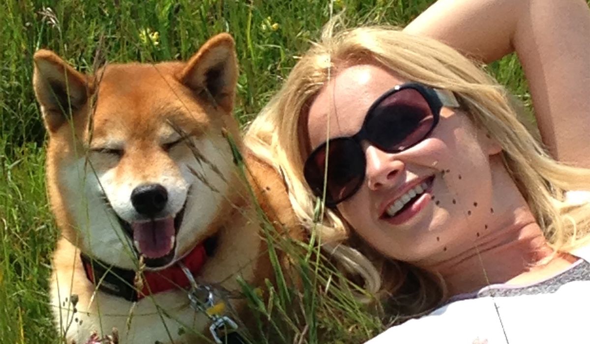 A Shiba Inu is lying in the grass with a lady, both are smiling in the sunshine
