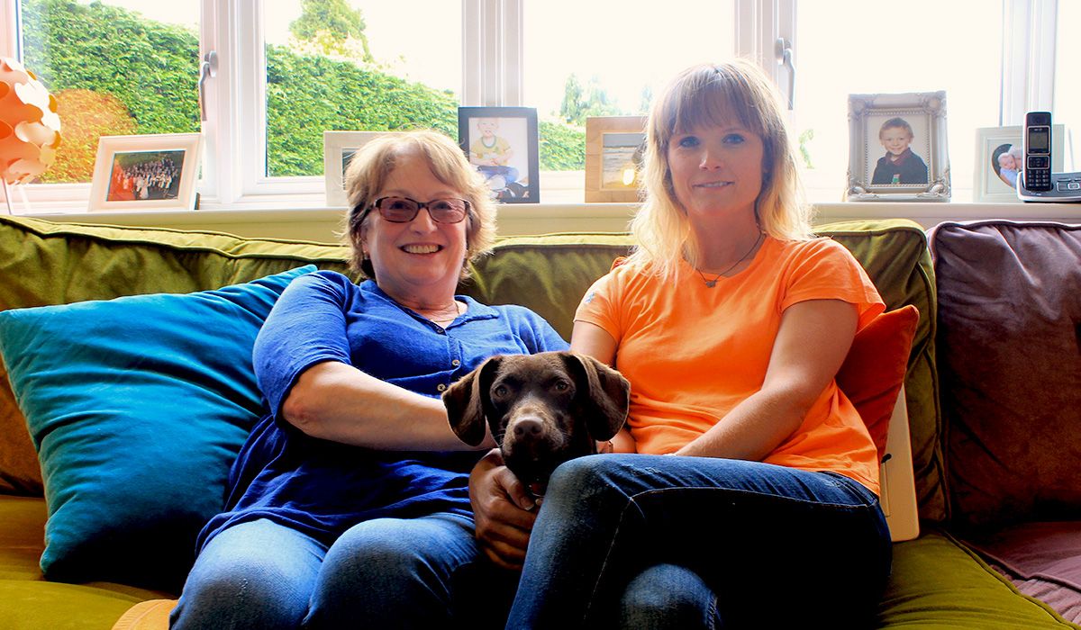 Two women are sitting on a sofa - on their laps is a small, short-coated brown dog who is looking intently at the camera