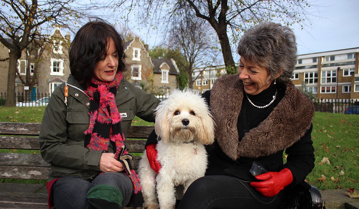 Two ladies are sitting on a bench, between them is with a small, white doggy