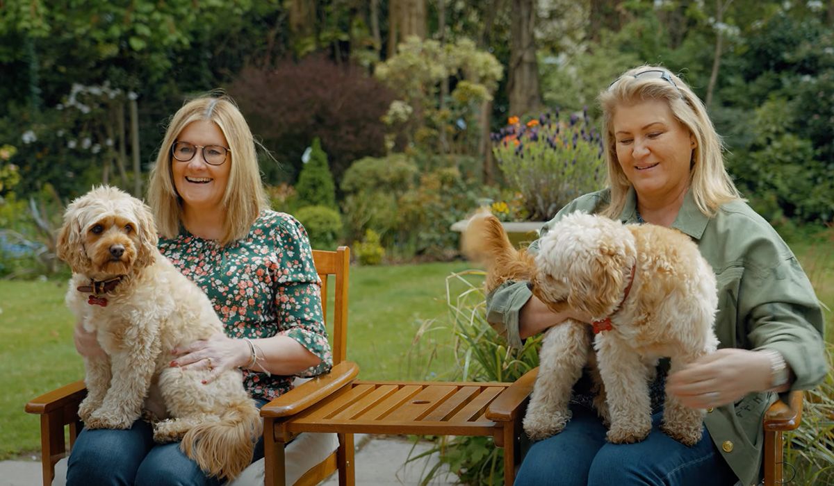 Two smiling women sit on chairs in a garden - each has a golden, curly-haired dog on their lap